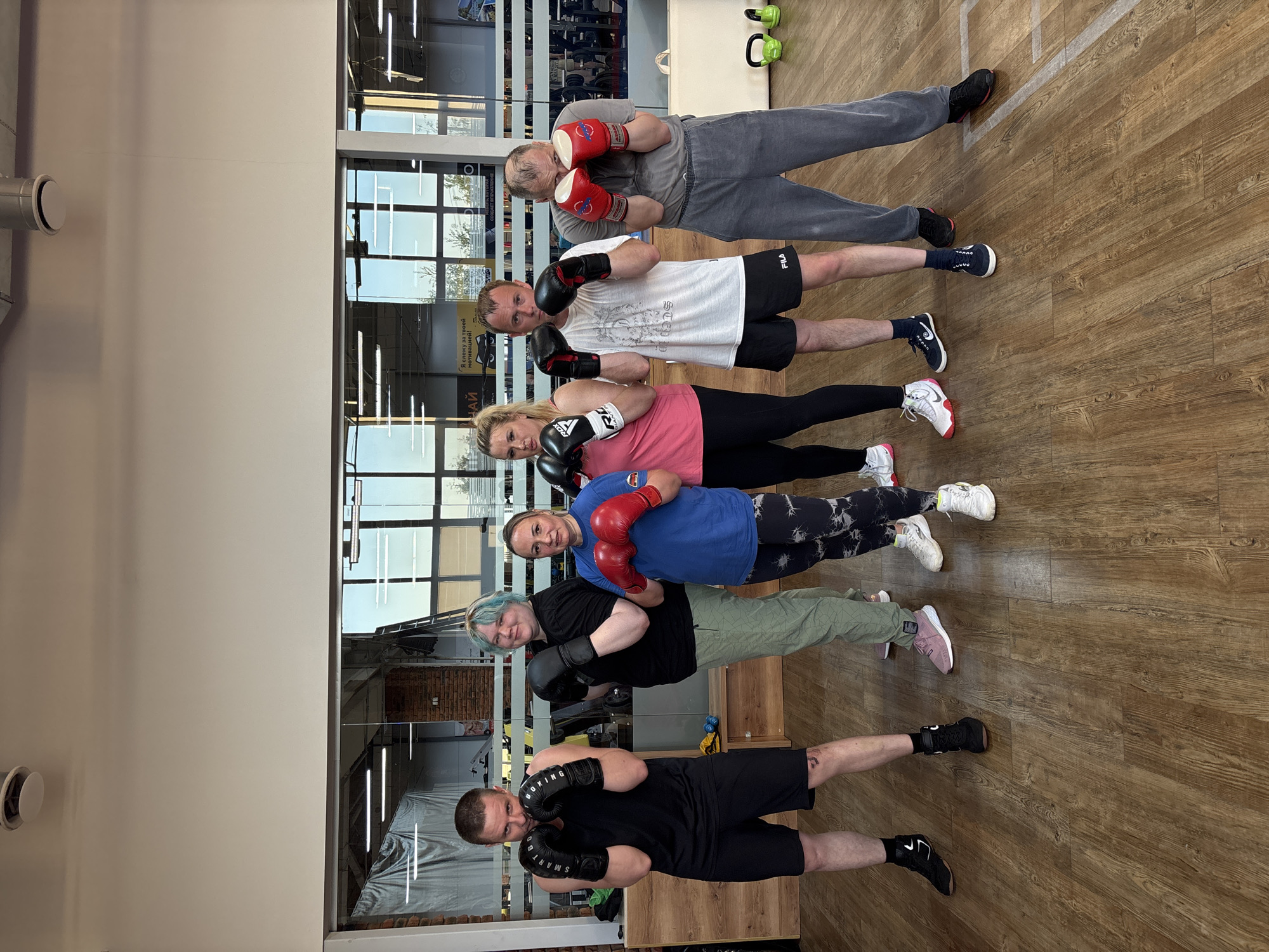 A group of six people stands in a line in what appears to be a gym or fitness studio. They are all wearing boxing gloves, some red and some black, and are holding them up in a defensive boxing stance. The group consists of both men and women of varying ages. They are dressed in casual athletic wear, including shorts, leggings, t-shirts, and sweatshirts. The gym has a wooden floor and large windows that reveal an exterior scene, including what seems to be a building with multiple windows and possibly some trees. There are gym equipment visible, including kettlebells in the background. Based on the architectural style of the building seen through the window, this image might be taken in a gym or fitness studio in the city of Chaikovsky, Russia.