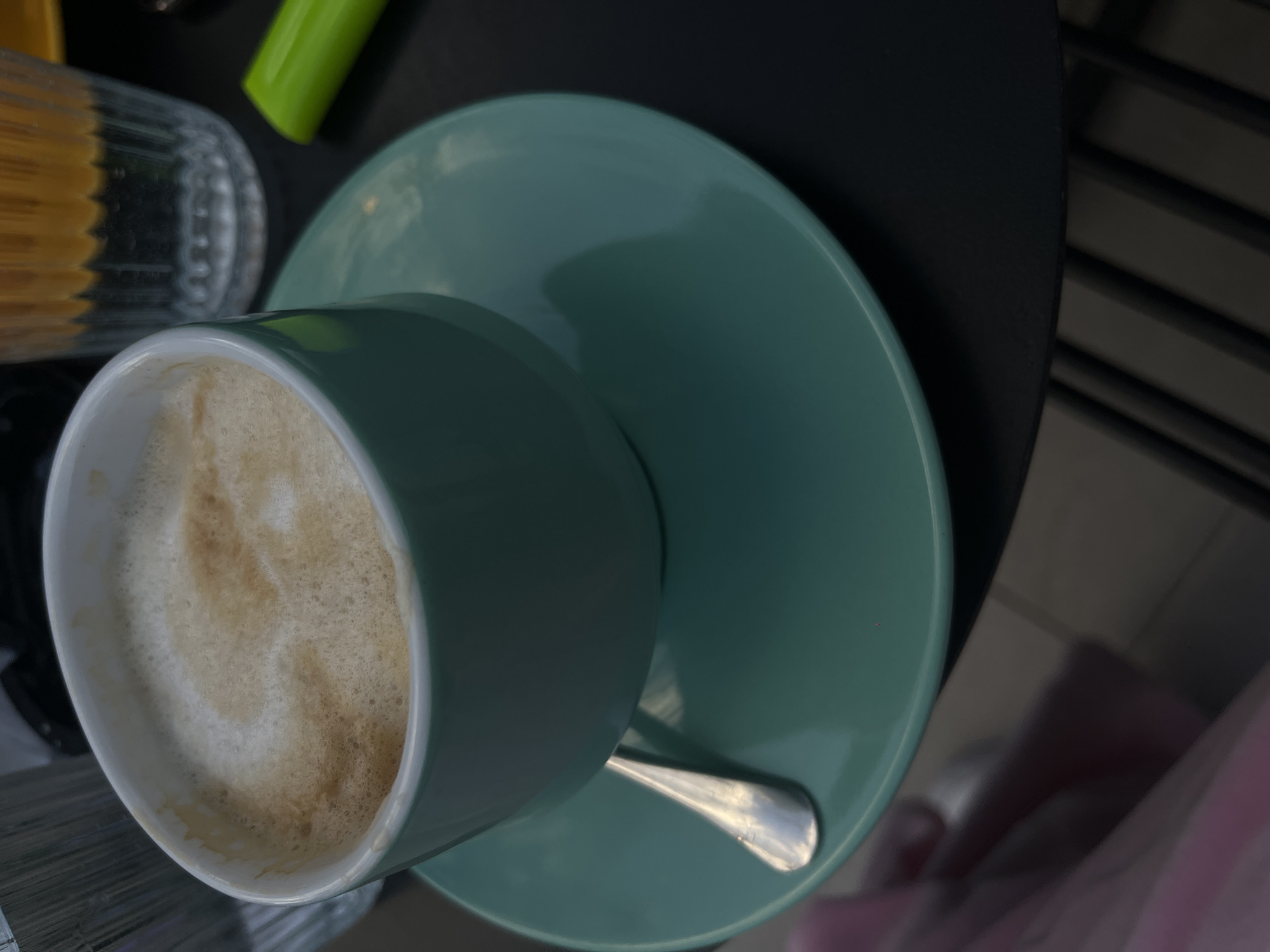 A high-angle shot shows a light green coffee cup filled with a beverage that has a frothy top, resting on a light green saucer. A silver spoon is placed on the saucer next to the cup. The saucer and cup sit on a black table or surface. In the background to the left, a partially visible glass container is present along with a blurred green object. A metal railing is partially visible on the right, with a hint of pink fabric near the railing.
The location is undetermined.