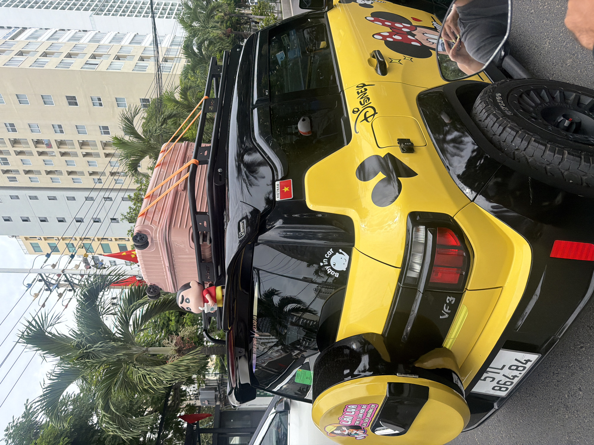 A yellow SUV with black accents is parked on a street in Nha Trang, Vietnam. The car is decorated with various decals, including Disney characters like Mickey and Minnie Mouse, and the word "Disney". A pink suitcase is secured to the roof rack. A small figurine of Shin-chan, a popular anime character, is positioned on the roof rack next to the suitcase. The license plate reads "51L 864.84". In the background, tall buildings and palm trees are visible, indicating an urban environment. The image appears to be taken from another vehicle or from ground level, focusing on the rear and side of the SUV.