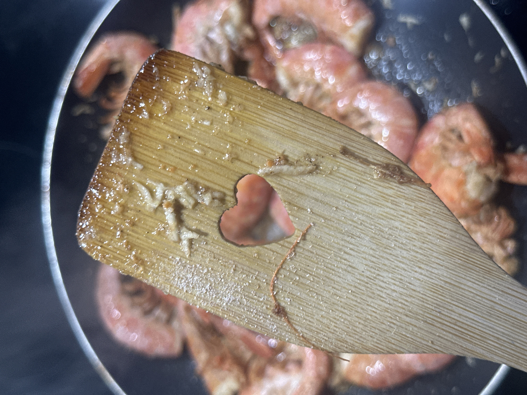 A close-up shot shows a wooden spatula with a heart-shaped cutout, coated with oil or food residue, positioned over a pan of cooked shrimp. The shrimp are pink and scattered within the pan. The edge of the pan is visible. The photo gives no indication of the location.