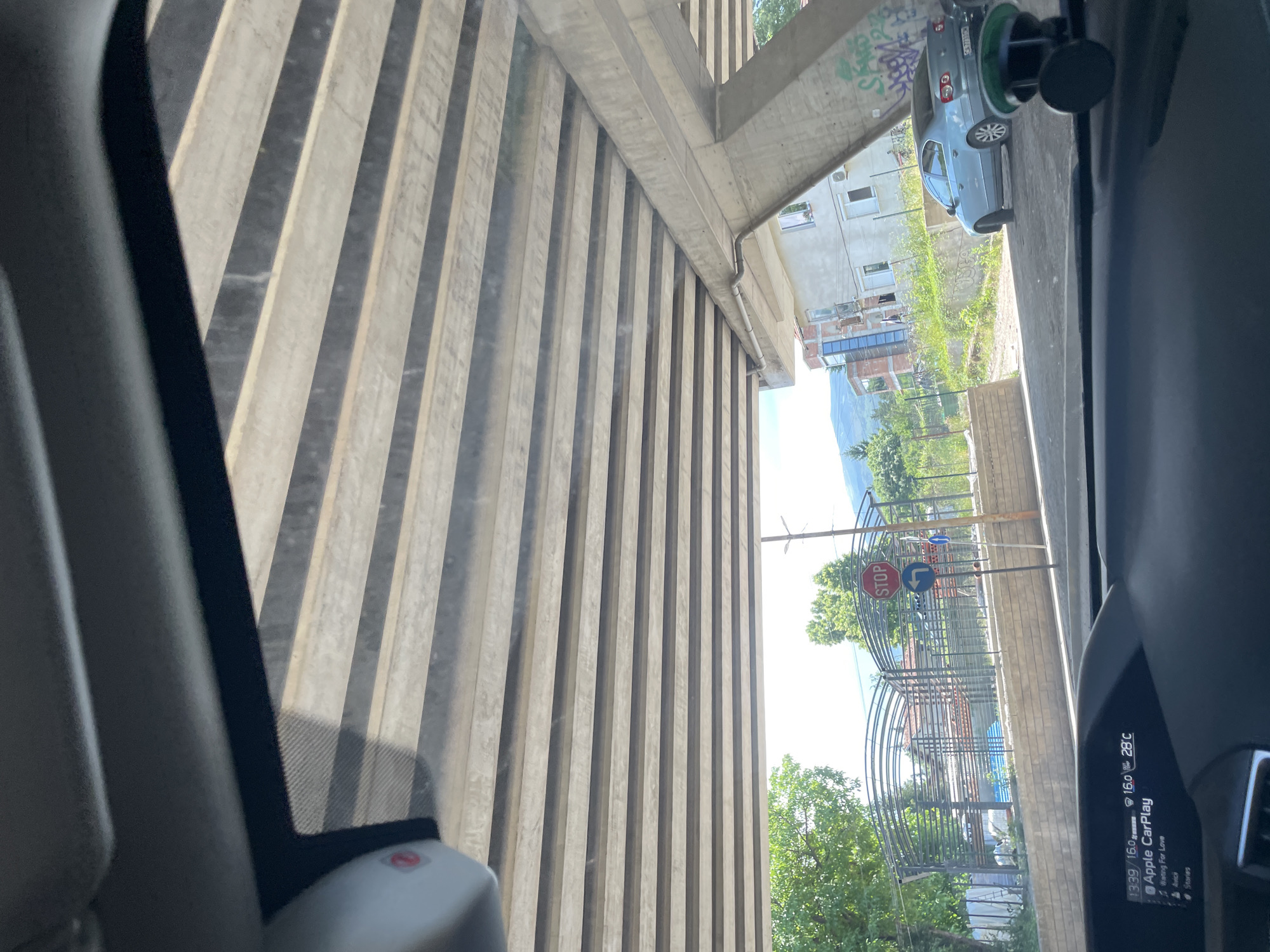 The image, taken from inside a car in Sofia, Bulgaria, shows a view of the underside of an elevated concrete structure, likely a highway or bridge. Sunlight streams through the gaps between the parallel concrete beams of the structure, creating a striped pattern of light and shadow across the scene.
In the distance, beyond a low wall, there are two buildings. The building on the left appears to be a modern apartment complex with a glass facade, while the building on the right is a more traditional, lighter-colored building with windows. Adjacent to the buildings are trees and vegetation. A light blue sedan is parked on the road to the right.
A red octagonal "STOP" sign is visible on a pole in the middle ground, accompanied by a blue directional arrow sign. The dashboard of the car is in the foreground, displaying the time as 13:39, a temperature of 28°C, and "Apple CarPlay" as the active interface, currently playing "Waiting For Love."