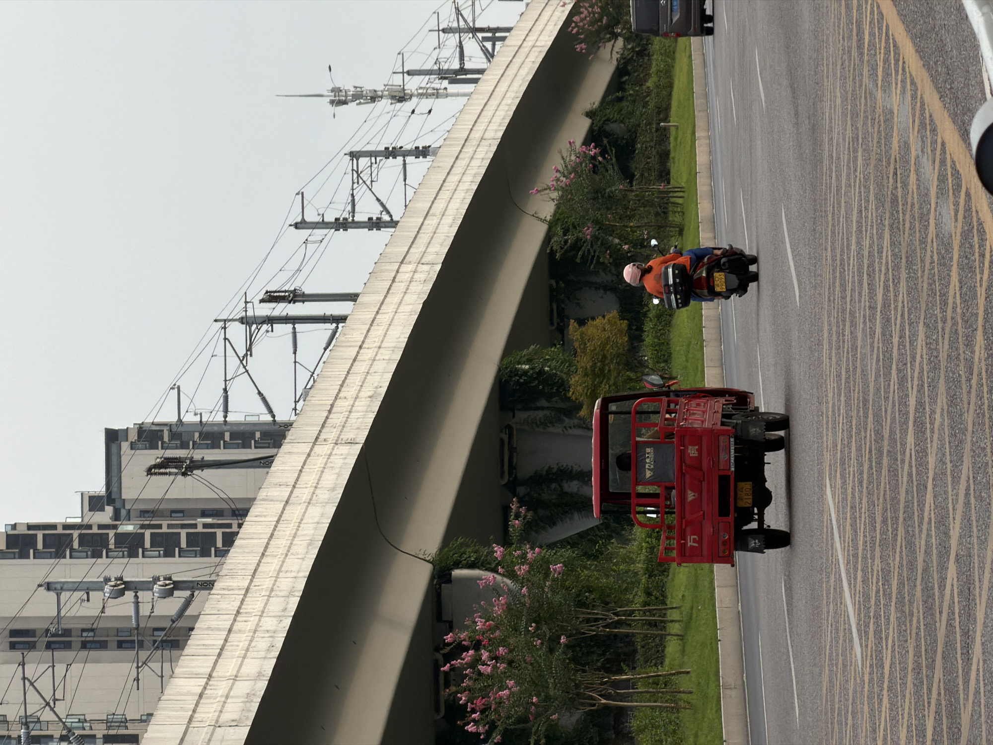 The image depicts a street scene in Jiangjin, China, with a focus on transportation infrastructure and vehicles. In the foreground, a red three-wheeled vehicle is traveling on the road. Behind it, a person on a scooter with an orange jacket and a pink helmet is also moving forward. A dark-colored vehicle is visible further down the road on the right. The background is dominated by a large elevated concrete structure, likely a bridge or overpass, with numerous electrical wires and poles extending from it. Behind the elevated structure, several tall, modern buildings are visible, suggesting an urban environment. Greenery, including trees with pink blossoms, lines the side of the road and grows beneath the elevated structure. The sky is overcast.