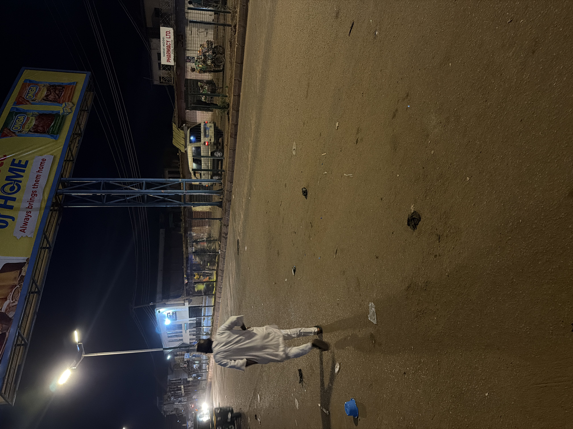 In Bukuru, Nigeria, at night, a man in white attire walks away from the camera on a wet asphalt road. The road is littered with debris, including plastic bags and a blue bucket. To the right of the road, a wire fence encloses a row of buildings and shops. A white van is parked in front of one of the buildings, and further down, a motorcycle is laden with goods. Above the scene, a large billboard displays advertisements for "OJ HOME" and "ONGA" products. A street lamp illuminates the area, casting long shadows.