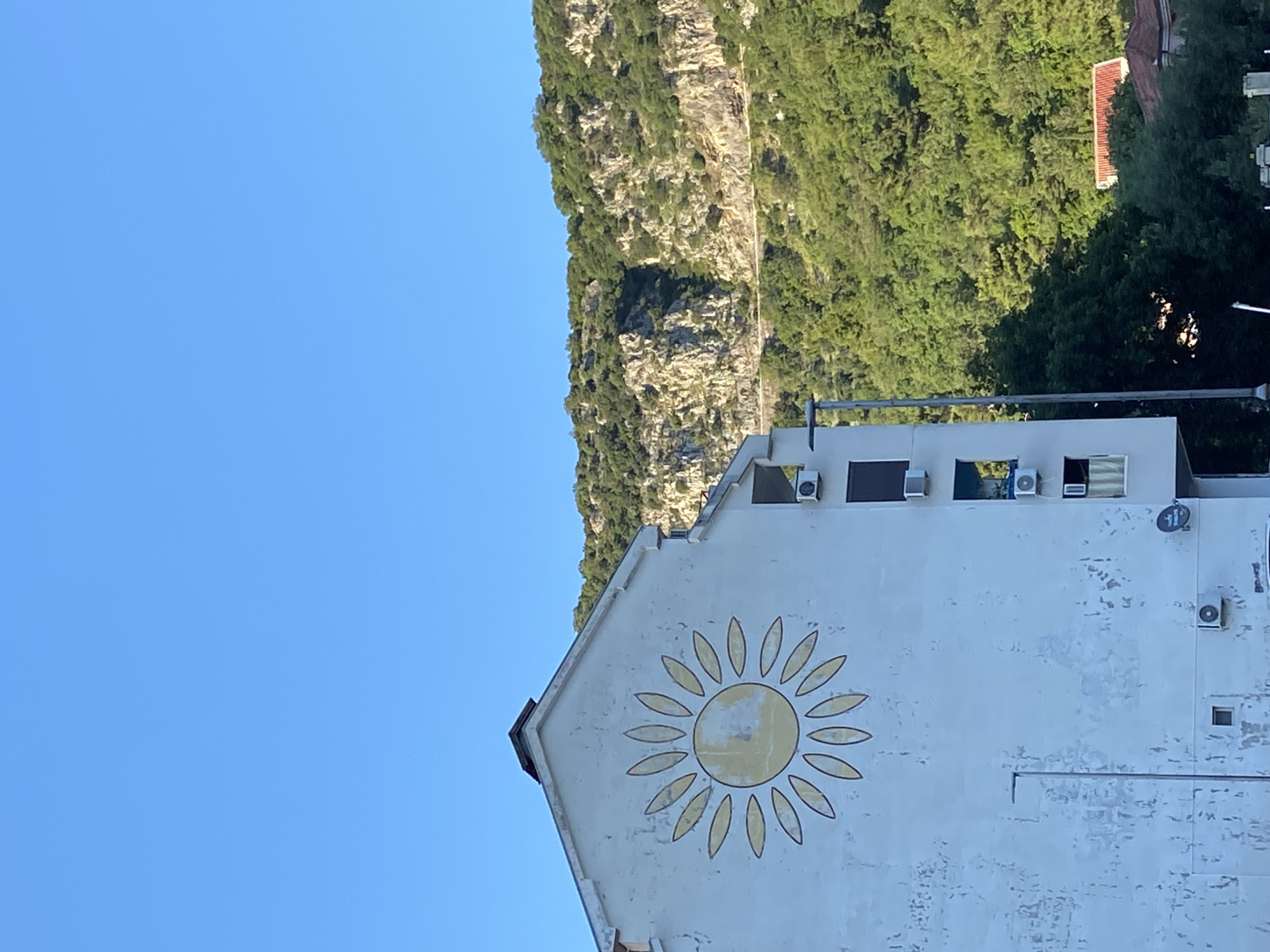 A building with a large sun painted on its gable stands in Budva, Montenegro, under a clear blue sky. A mountain covered in green vegetation rises in the background, partially obscuring the building's upper floors. Multiple air conditioning units are attached to the building's facade.