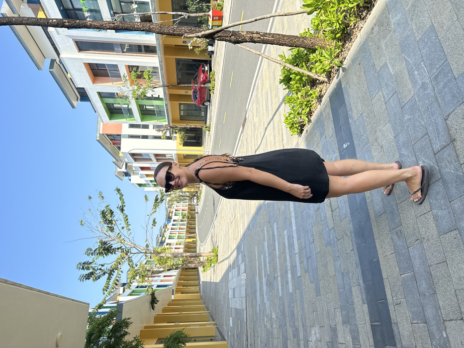 The image depicts a solitary female subject standing on a grey rectangular tiled sidewalk in an urban development in Phú Quốc, Vietnam. The subject, with dark hair pulled back, wears dark-framed sunglasses, a black knee-length sleeveless dress with visible leopard-print patterned straps on the upper back, and dark-colored flip-flops. Her body is angled slightly to the right, with her head turned over her left shoulder, directly facing the viewer and exhibiting a smile.
The background features a clear blue sky and a street lined with modern, multi-story buildings characterized by contemporary architecture and facades painted in varying vibrant colors, including yellow, white, green, orange, blue, and red. A paved road runs through the center of the scene, with a single red car parked parallel to the curb on the right side. Sparse deciduous trees and low green shrubs are planted along the sidewalk and street. Long, defined shadows are cast on the sidewalk and street, indicating bright, direct sunlight. No other individuals or interactions are visible within the frame.