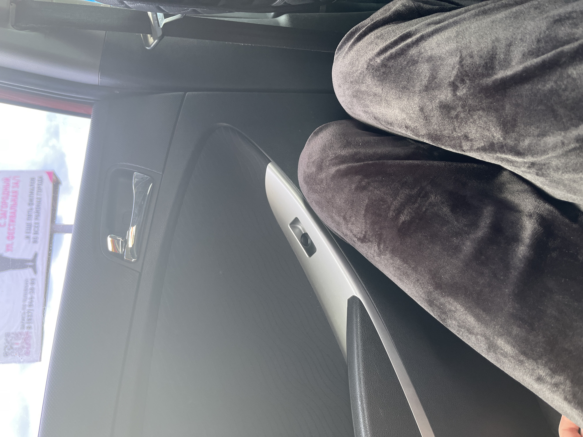 The image shows the interior of a car. In the foreground, a person's legs, clad in dark gray velour pants, are visible. The pants are creased, indicating the person is sitting. The car's interior includes a black door panel with a silver trim and a chrome door handle. A silver-colored window control panel is visible on the door. Beyond the car's interior, a blurry billboard with text in Russian is visible through the car window, suggesting an outdoor urban or suburban environment. The billboard appears to have a QR code and phone numbers. The lighting suggests it is daytime. The location is identified as Novaya Otradovka, Russia.