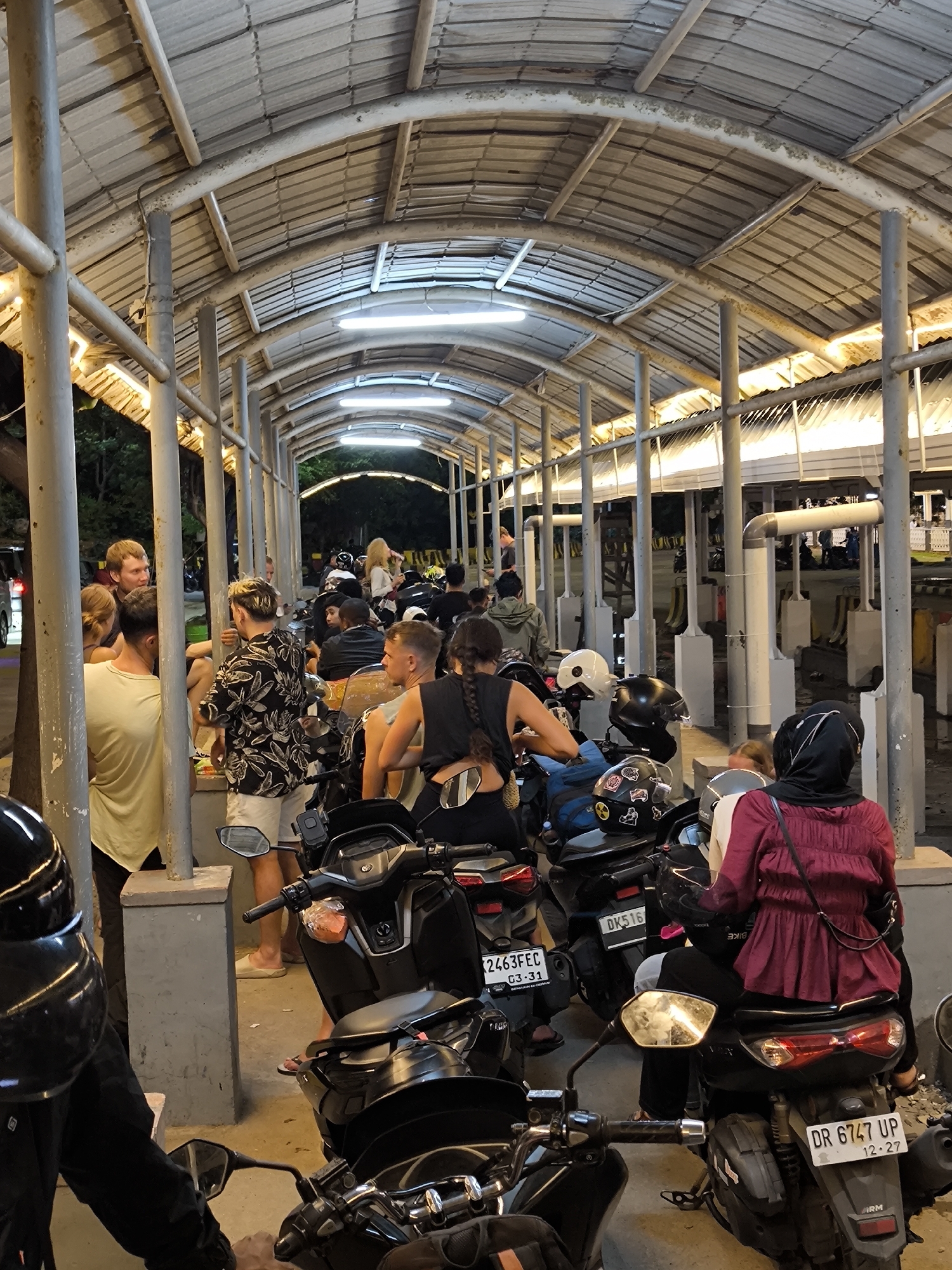 Here is a detailed description of the image:
Location: The image shows a bustling scene in what appears to be an outdoor food market in Unknown, Unknown.
People: There is a diverse group of people, likely customers, waiting in a queue. Many are standing and interacting with each other, while others are seated on their parked scooters.
Objects: The prominent objects in the image are the scooters, which are parked in an orderly manner. The scooters are primarily black, with varying license plates. There is a covered walkway with metal support beams and fluorescent lighting, offering shelter from the elements. The walkway also features a series of individual food stalls, identified by the metal framing around them.
Actions: The primary action is people waiting in line, likely to purchase food or drinks. Some individuals are engaged in conversations, while others are focused on their scooters.
Interactions: The interactions observed involve people talking to each other, sitting on their scooters, and generally waiting their turn.
Environment: The environment suggests a vibrant night market atmosphere. The lighting is warm, providing a sense of activity and commerce. The area appears to be relatively crowded, indicating a popular destination for both locals and tourists.