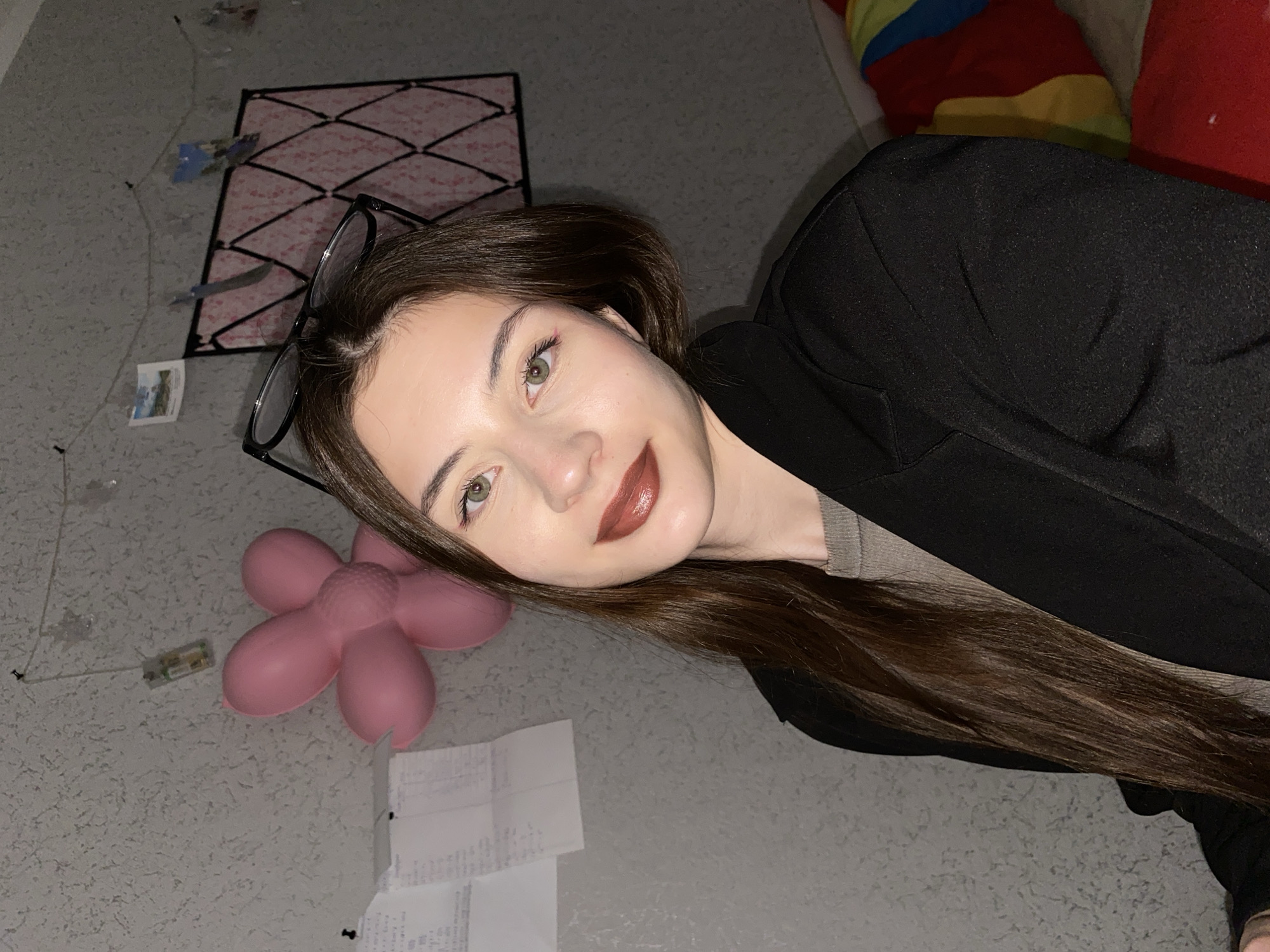 A young woman with long, dark brown hair and light green eyes is captured in a close-up, rotated portrait, likely a selfie, in an indoor setting in Votkinsk, Russia. She is looking directly at the camera with a subtle smile, her lips adorned with dark, glossy lipstick. Her dark-rimmed glasses rest on her head. She wears a dark, possibly black, jacket or blazer over a lighter, perhaps grey or olive-green, top.
The background suggests a casual room. The dominant surface, likely a floor or wall, is light grey with a textured finish. To her left (viewer's left), a large, pink, flower-shaped object, possibly a cushion or inflatable toy, is visible. Near it, a small electronic component with thin wires is present. Further to the left, crumpled or folded white papers, resembling receipts, are scattered, with one held down by a thumbtack.
Above her head, a black grid structure with a pink backing is pinned to the wall, possibly serving as a memo board or a decorative item, with small pictures or notes attached to it. In the upper right corner of the frame, a colorful, striped pillow or blanket with red, yellow, and blue sections suggests a soft furnishing like a bed or sofa. The lighting is soft and appears to be artificial indoor light. No discernible text is visible on the scattered papers or other objects due to the distance and resolution.