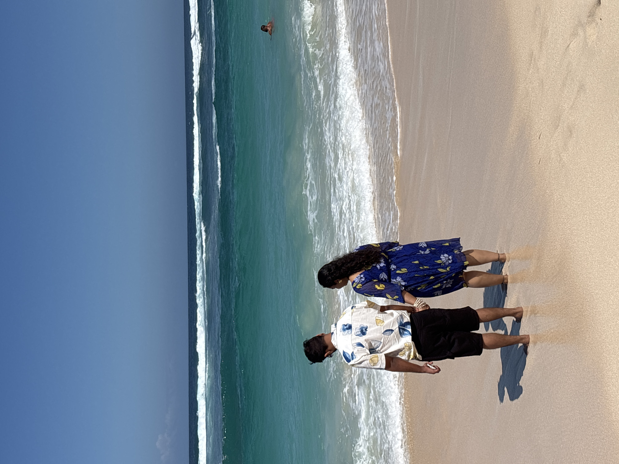 A rear-view medium shot captures a couple standing on a sandy beach in Kuta Selatan, Indonesia, holding hands and facing the turquoise ocean under a clear blue sky. The man, on the left, wears a white short-sleeved shirt with a blue floral pattern and black cargo shorts. The woman, on the right, is dressed in a blue floral-patterned dress. Their feet are submerged in shallow water at the edge of the shoreline. In the distance, waves are breaking, and a single person can be seen swimming in the ocean. The sand is light brown and appears wet where the waves reach it, with shadows cast by the couple.