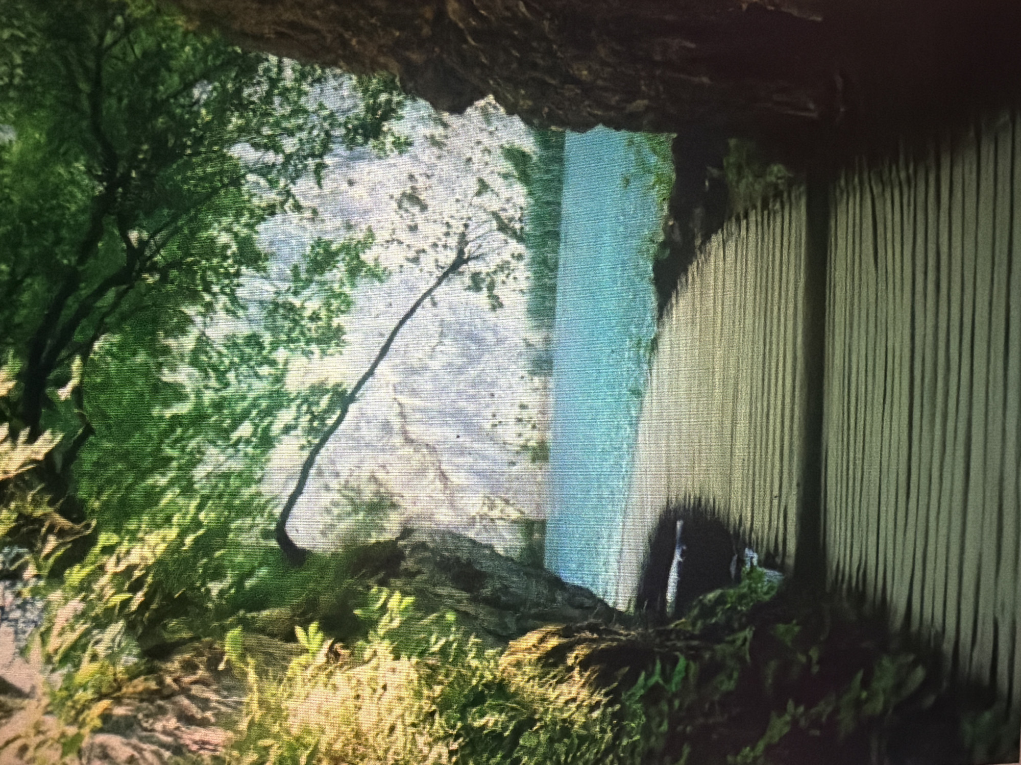 A wooden boardwalk curves into a body of turquoise water, framed by lush green foliage and the rugged texture of a cliff face. The scene is bathed in natural light, with dappled shadows cast by overhanging tree branches. The boardwalk is constructed of weathered planks laid horizontally, leading the viewer's eye towards the serene water and the distant, rocky landscape. The environment suggests a natural park or a scenic pathway, possibly near a lake or a calm sea inlet. There are no people or animals visible in the image. The location is identified as Baan Mae Nam, Thailand.