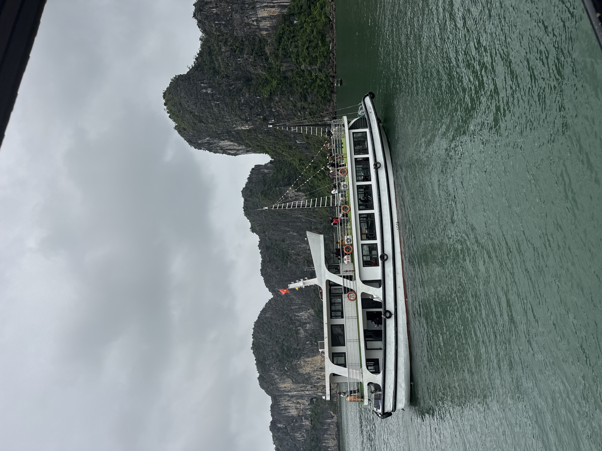 The image depicts a tour boat navigating through the emerald waters of Ha Long Bay, Vietnam. Towering karst formations covered in lush greenery rise dramatically from the sea, creating a breathtaking natural landscape. The sky is overcast with a soft, diffused light, contributing to the moody atmosphere.
The tour boat, painted white with a red waterline, has multiple decks and is filled with passengers enjoying the scenery. People are visible on the upper deck, some standing and looking out at the bay, while others are seated. Life rings are strategically placed along the railings for safety. A Vietnamese flag flies proudly from the mast of the boat.
In the background, other karst islands are visible, some appearing closer and more detailed than others. The water's surface is slightly rippled, reflecting the muted colors of the sky and the surrounding landscape. The scene captures a moment of leisure and exploration in one of Vietnam's most iconic natural wonders.