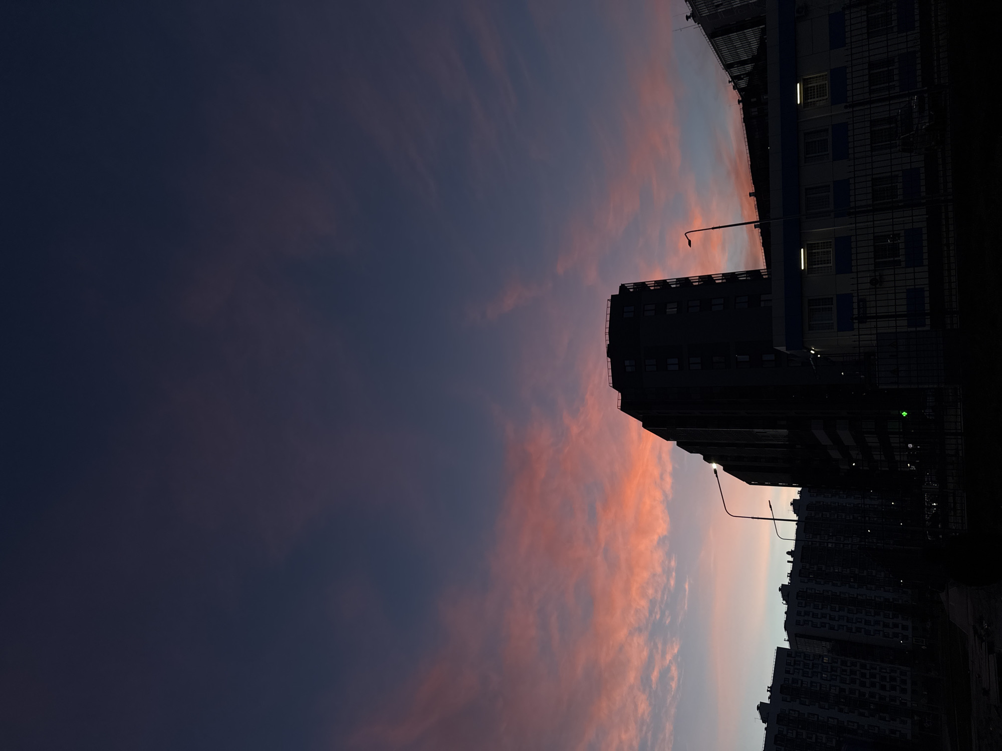 The image displays an urban twilight scene, rotated 90 degrees clockwise from a typical landscape orientation. The left and upper portions are filled with a striking sky that transitions from a deep, dark purplish-blue in the upper left to soft pinks and oranges in the lower right, eventually fading into a lighter blue on the far right. Wispy clouds, tinged with the sunset or sunrise colors, are scattered across the brighter parts of the sky.
Along the right side and wrapping towards the bottom, a series of multi-story buildings stand in silhouette. A prominent, dark building occupies the upper right side, with faint internal lights visible through some windows. Below this, another building features a light blue-grey facade with numerous rectangular windows, several of which are distinctly illuminated with a yellow-white light. Further right and along the bottom edge, a dense cluster of additional dark, silhouetted buildings forms an urban skyline.
Two streetlights are visible on the right side, extending horizontally from the buildings towards the sky, their light sources either dim or silhouetted. The dramatic sky colors and the artificial illumination from the buildings indicate that the time of day is twilight, either dusk or dawn. No specific activities or people are visible, and there is no discernible text. The scene presents a tranquil view of an urban environment, fitting the provided context of an unknown city in Russia.