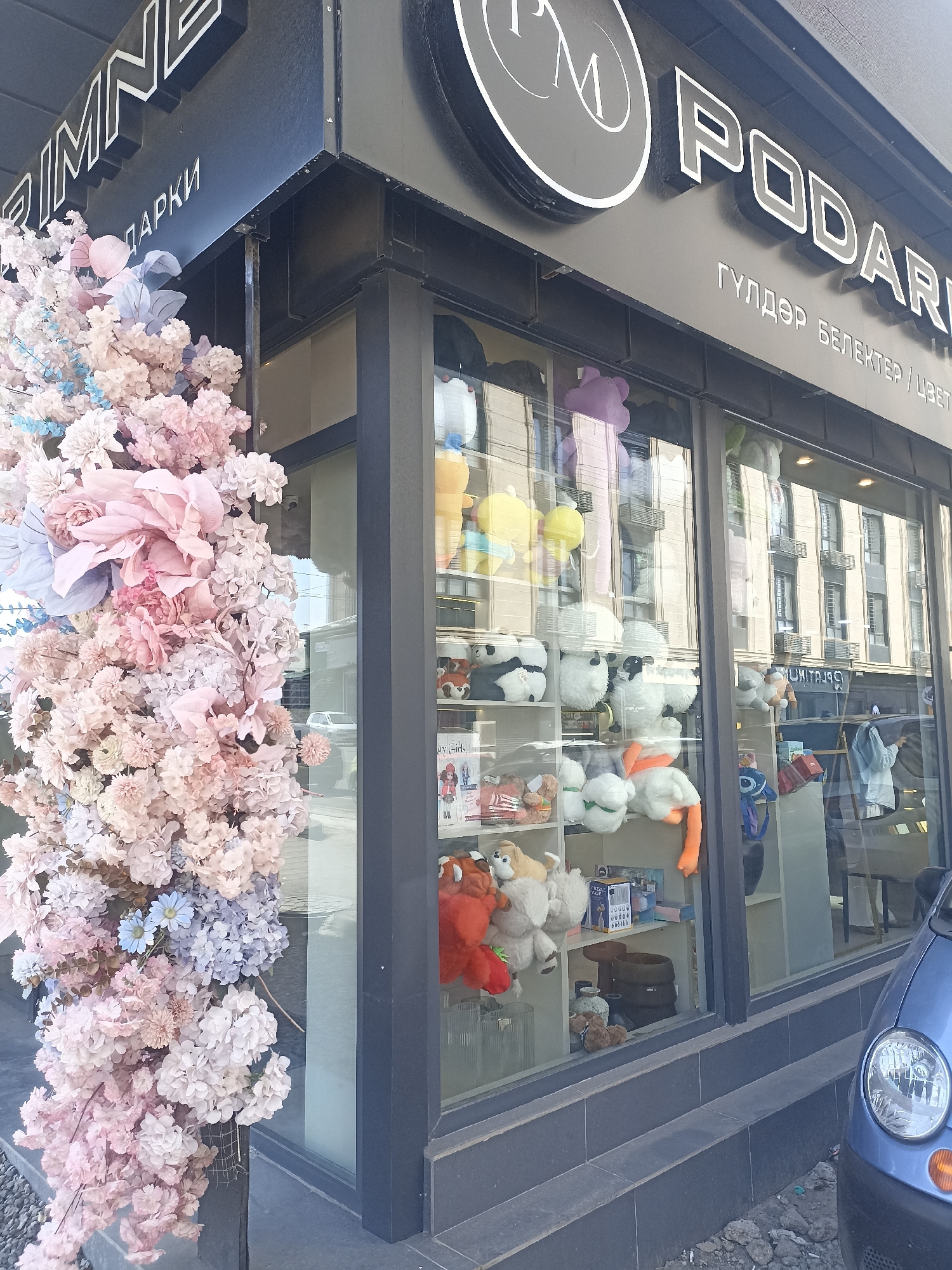 A storefront in Osh City, Kyrgyzstan, is decorated with large, pastel-colored artificial floral arrangements. The shop's sign, written in a stylized font, reads "PODARI" in large white letters against a black background. Below the main sign, smaller text in a silver font says "ГУЛДЭР БЕЛЕКТЕР / ЦВЕТ" which translates to "Flowers Gifts / Color". The shop window displays an assortment of stuffed animals, including teddy bears, pandas, and other characters. A blue car is parked to the right of the storefront. The street in front of the shop is paved with grey stones and gravel, and a concrete sidewalk is visible.