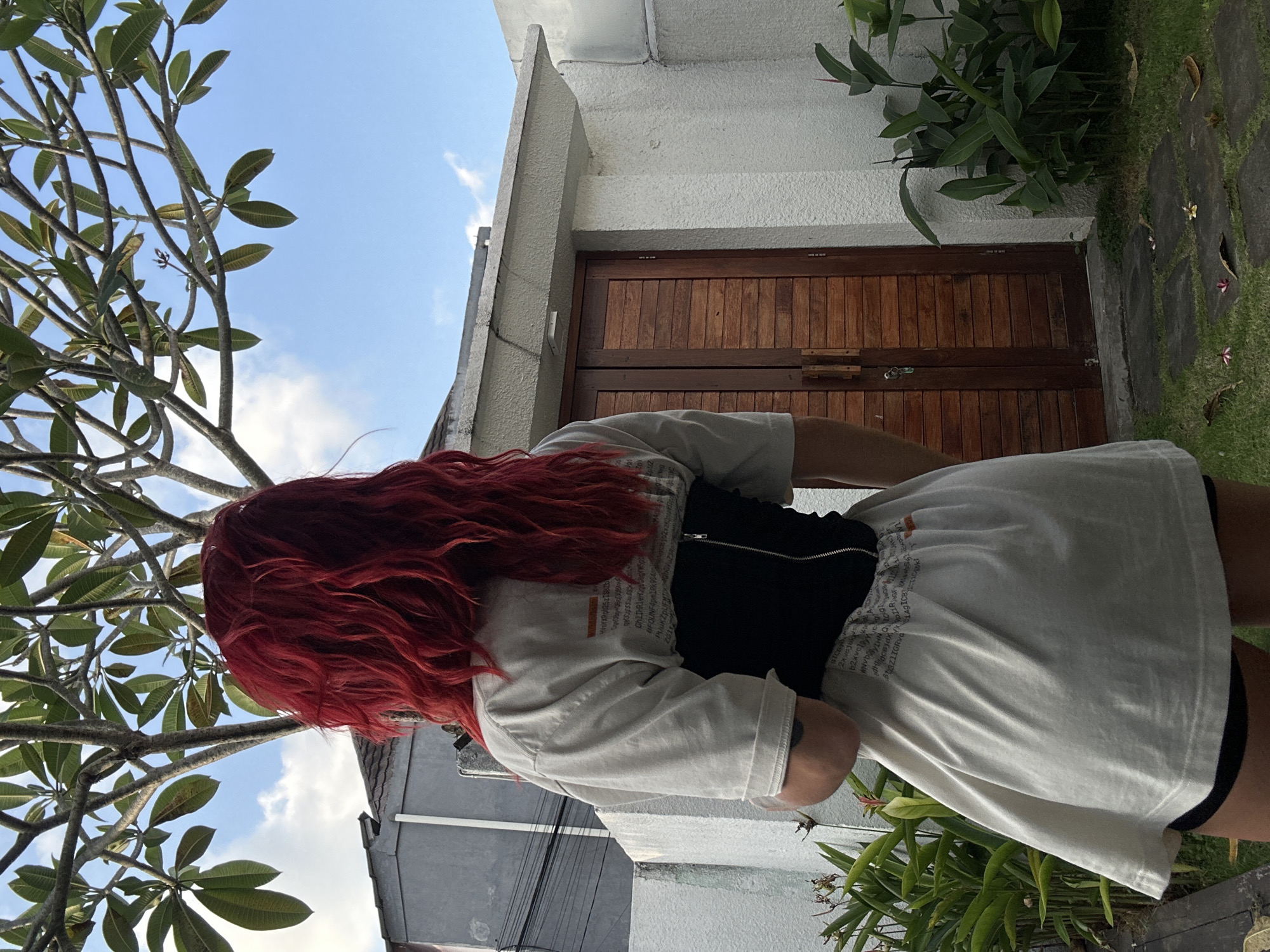 An individual with long, vibrant red hair is observed from behind, standing outdoors in North Kuta, Indonesia. The person wears an oversized light-colored t-shirt featuring dark text print on the back, cinched at the waist by a black corset with a visible silver zipper running vertically down the center back. Black shorts are visible beneath the t-shirt hem. The subject faces away from the viewer, looking towards a wooden double door set into a textured white building wall. Above the door, a light-colored concrete overhang extends. To the left, a grey building with a dark roof is partially visible. The immediate ground features grass with interspersed grey stone pavers and various green leafy plants. Overhead, a large tree with numerous branches and dense green leaves (likely a Plumeria or Frangipani) fills the upper portion of the frame, silhouetted against a clear blue sky with sparse white clouds.