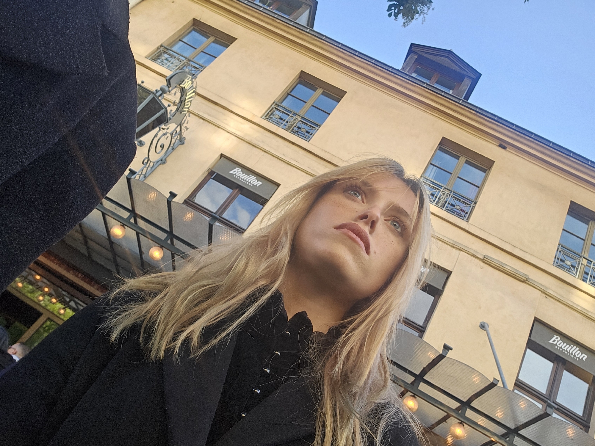 The photograph depicts a woman with blonde hair, wearing a black coat and top, looking upwards. The shot is taken from a low angle, capturing a building in the background. The building has multiple windows with dark frames and a sign that reads "Bouillon." The architectural style suggests the location is in Paris, France. Metal bars and lights are visible above the woman, likely part of an outdoor seating area or awning. A small portion of a person's head is seen blurred in the lower left corner. The sky above the building appears light blue.