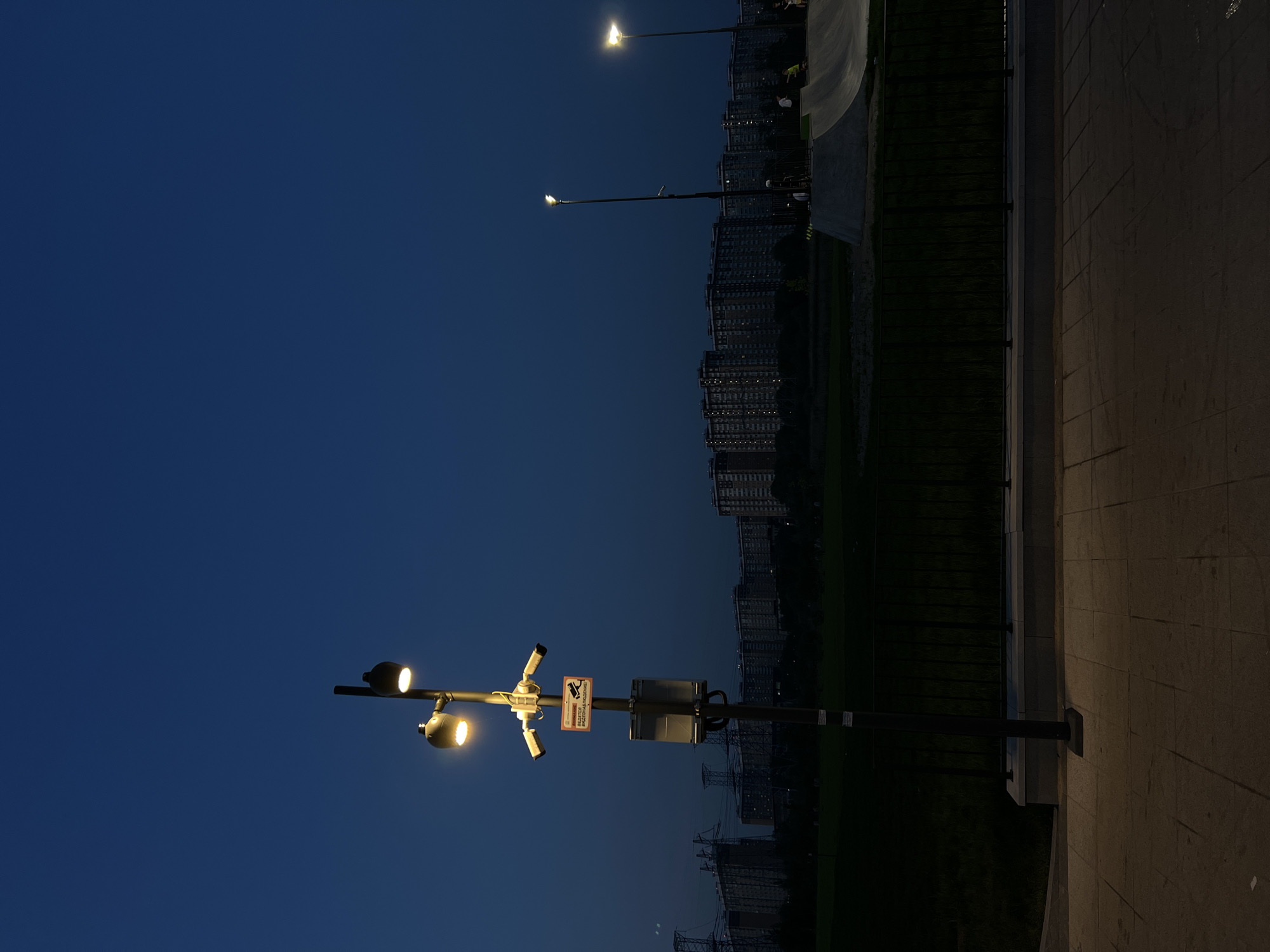 The image depicts an urban scene at night in Moscow, Russia. A modern streetlight post with attached surveillance cameras and a sign is visible in the foreground. In the background, rows of high-rise apartment buildings are illuminated, indicating a densely populated residential area. Further in the background is a lake and green space between the apartment buildings. The sky is a dark blue. The foreground shows concrete tiles.