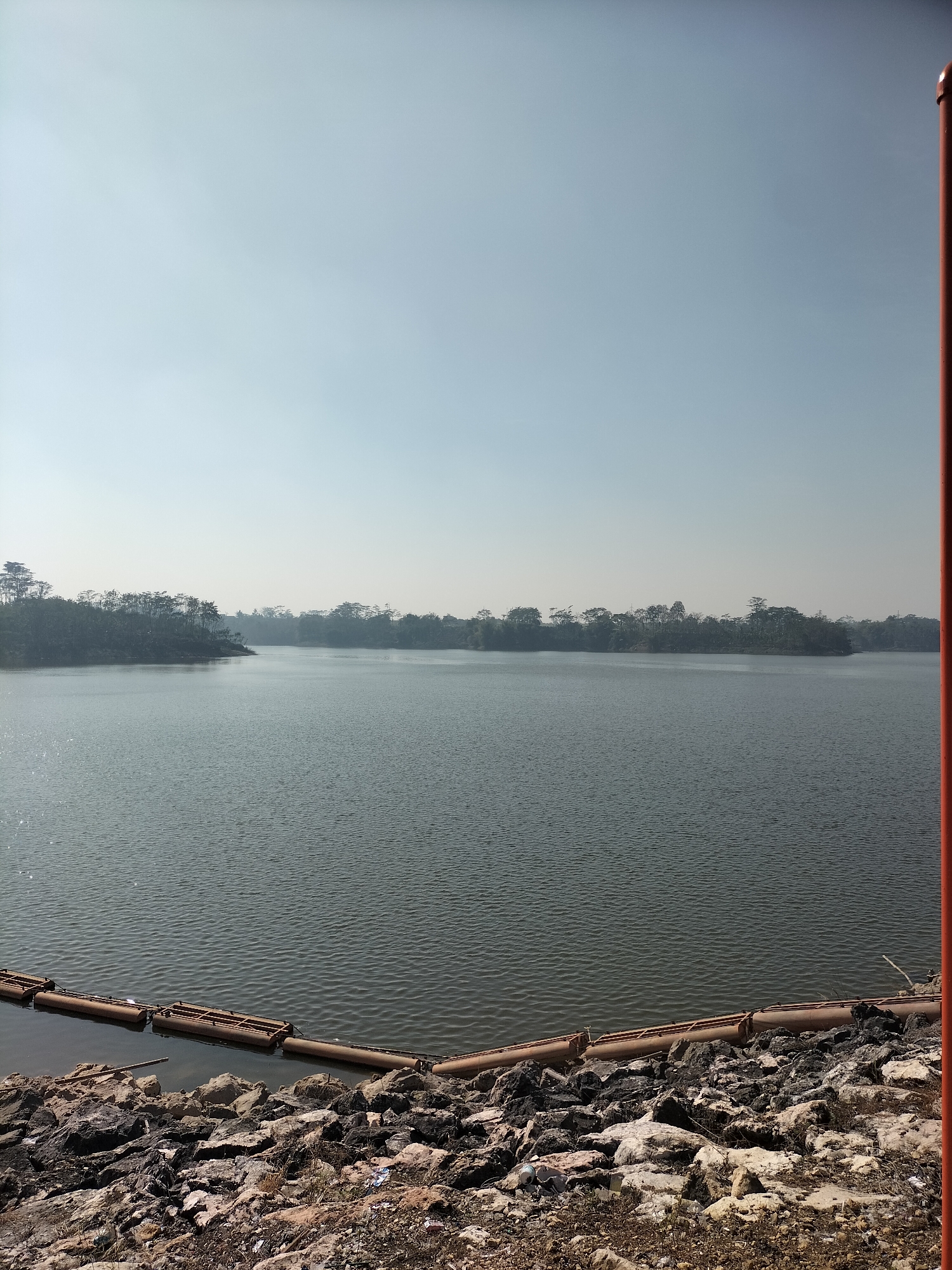 The image depicts a serene reservoir in Sumberpucung, Indonesia. A calm body of water dominates the scene, reflecting the clear, pale blue sky. The banks of the reservoir are lined with rough, grey rocks and sparse vegetation. In the foreground, a series of connected, rusty-orange pipes float on the water's surface, possibly serving as a boom or barrier. The distant shoreline is a dense line of trees, their dark green foliage contrasting with the lighter sky. A vertical, reddish-orange pole is visible on the far right edge of the frame. The overall atmosphere is tranquil and natural, with no people or animals present.