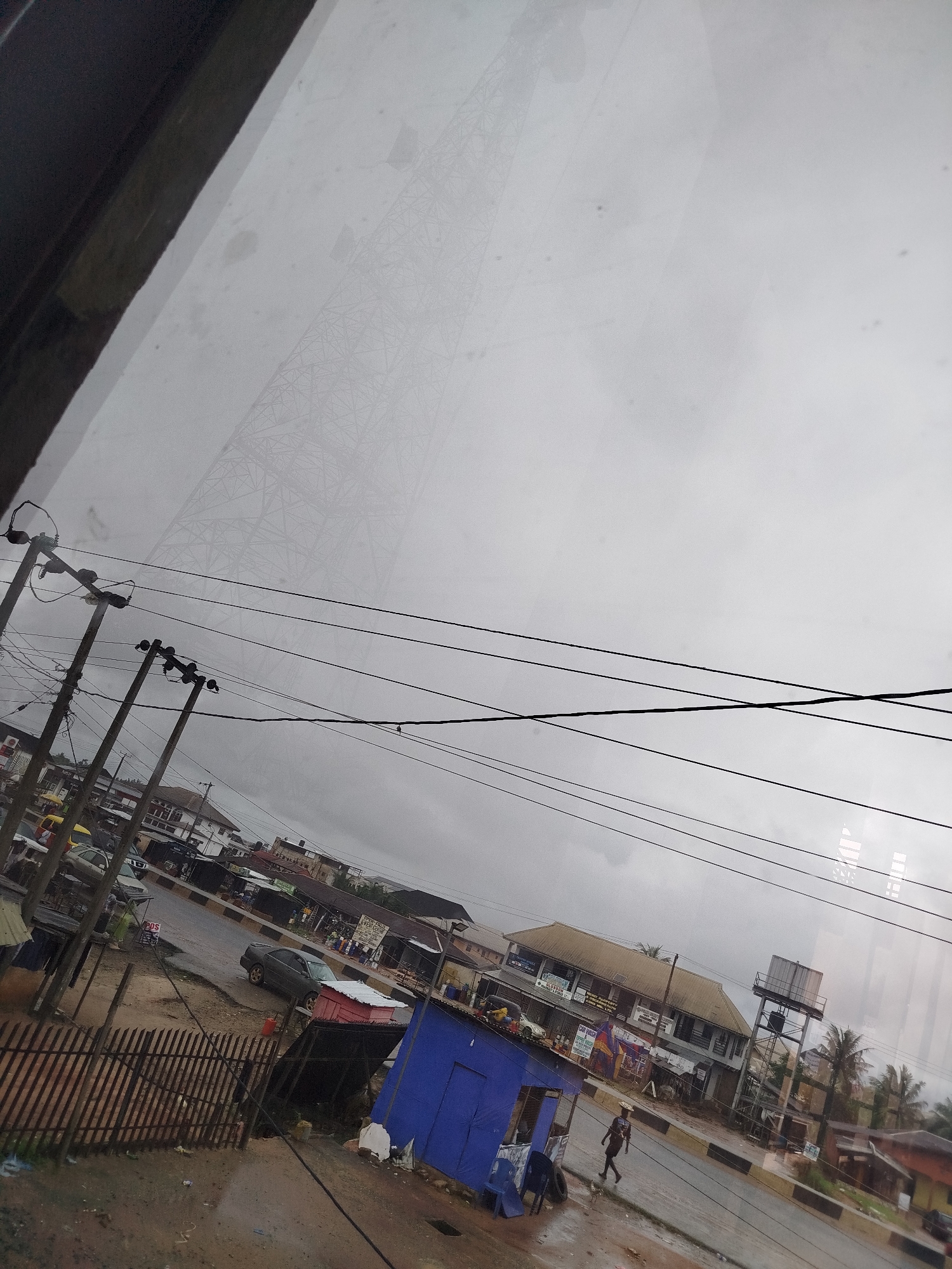 The image depicts a street scene in Uselu, Nigeria, under an overcast sky. The perspective is from an elevated position, possibly looking through a window. In the foreground, a wooden fence runs along the left side of the image, partially obscuring a dirt and gravel path.
Dominating the middle ground are utility poles with multiple electrical wires stretching across the frame. A dark-colored sedan is parked on the wet asphalt road. To the right of the road, a small, bright blue building with a red-tiled roof is visible. Further back, several other buildings line the street, with awnings and signs suggesting commercial activity. One building has a light-colored, tiled roof and multiple windows. A water tower with a large tank sits behind some of these structures. Palm trees are scattered throughout the scene, indicating a tropical or subtropical climate.
In the distance, a large lattice tower, likely a telecommunications tower, is faintly visible against the hazy sky. The overall impression is of a functional, somewhat weathered urban environment. A single individual, carrying an object on their head, walks along the sidewalk in the middle ground, adding a human element to the scene. The road appears damp, suggesting recent rain.