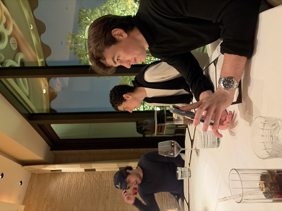 Three young men are seated at a table in a restaurant. One man, wearing a black shirt, is using a cell phone with a stainless steel wristwatch visible on his wrist. Another man, wearing a white shirt with a black jacket, is looking down, presumably at his own device. A third man, wearing a dark baseball cap and a dark shirt, appears to be on a phone call. The table is set with various glasses and what may be menus. The restaurant has a modern interior with a window view.