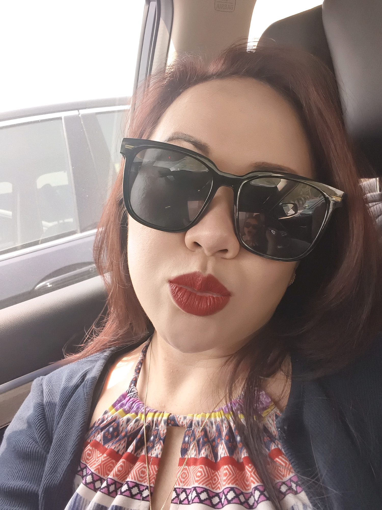 A woman with brown hair and red lipstick is in the passenger seat of a car. She is wearing large, dark sunglasses and a colorful, patterned top with a blue cardigan over it. The car's interior is visible, including the headrest and the window. The scene suggests an outdoor environment, possibly a street or a parking lot. There is no specific indication of the city being Dubai, United Arab Emirates.
