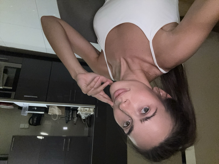 A woman poses indoors in a white tank top. She is fair-skinned, with long, dark hair. She holds her hand near her face. A modern kitchen is visible in the background with dark cabinets and appliances. The image is taken from a close-up angle and is slightly rotated.