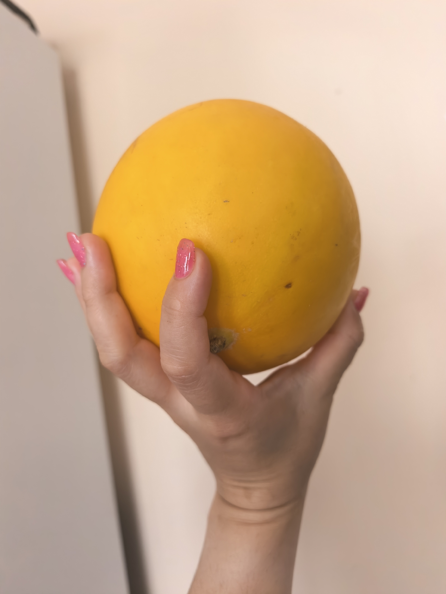 A close-up shot shows a hand holding a ripe cantaloupe. The hand, with bright pink, glitter-flecked fingernails, gently cradles the fruit. The cantaloupe is a vibrant yellow-orange, with a smooth rind that shows a few tiny brown speckles. A small bit of the stem attachment point is visible near the base of the fruit. The background is a soft, neutral tone, possibly a wall, with a blurred light-colored object on the left side of the frame. The lighting suggests an indoor setting, likely during the daytime. The overall impression is a simple, still-life composition highlighting the texture and color of the fruit and the polished appearance of the hand. There is no visible text in the image.