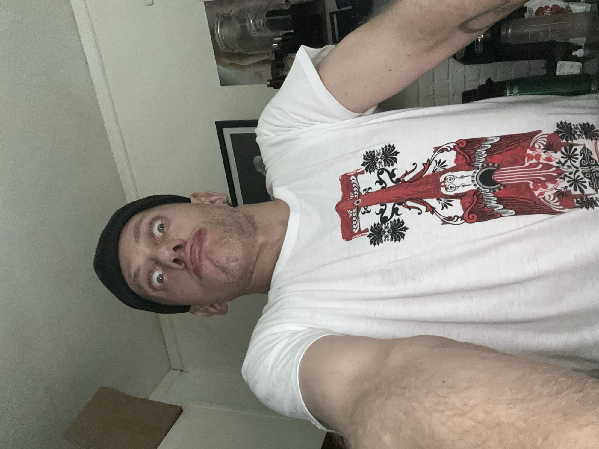 A man wearing a black beanie and a white t-shirt with a red and black graphic print is looking at the camera with a goofy expression. His eyes are wide and his mouth is pursed. The man's right arm is extended towards the camera, and his left arm is bent at the elbow. In the background, there is a white wall, a framed picture, and some bottles on a shelf. The scene appears to be indoors. There is no identifiable information to link the image to Moscow, Russia.