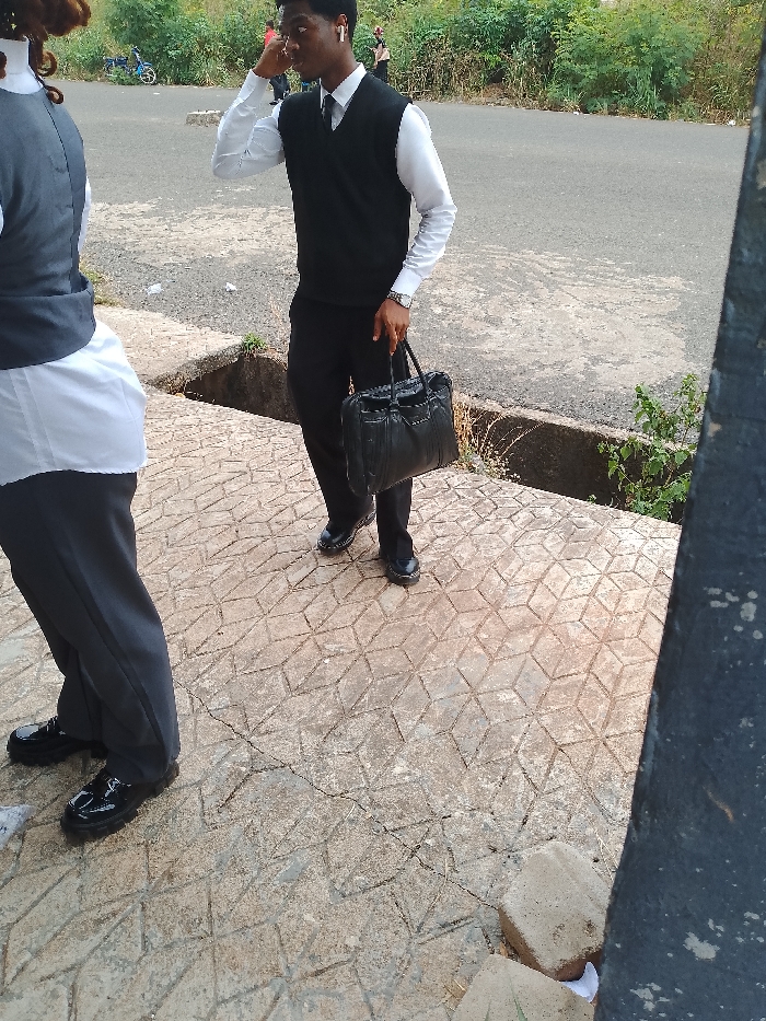 An outdoor scene in Ogboko, Nigeria, captures two individuals on a patterned concrete sidewalk. The primary subject, a male, stands on the right, wearing a white long-sleeved shirt, black tie, black V-neck vest, black trousers, and shiny black dress shoes. He has white in-ear headphones in both ears and a silver-toned wristwatch on his left wrist. His right hand is positioned near his right ear, and his left hand holds a black, soft-sided briefcase. To the left, a second individual is partially visible from the lower torso down, facing away from the camera, wearing a white shirt under a dark grey vest, dark grey trousers, and shiny black dress shoes. The concrete pavement, featuring a geometric pattern, shows signs of wear and minor cracking. An asphalt road, separated by a concrete drainage ditch, is visible behind the individuals. The background displays green foliage and bushes, with a blue bicycle faintly discernible on the distant road.