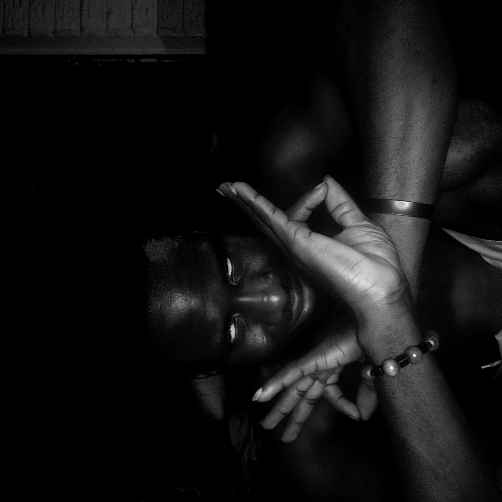 A close-up, black and white photograph shows a dark-skinned person with dark hair and visible eyes. The person is lying down and tilted on the side with their face towards the camera. The person is making a hand gesture, possibly a mudra. The person is wearing a bracelet of light-colored beads on one wrist and a cuff on the other. The background is dark and indistinct. The location in Narayi, Nigeria is not readily discernible.