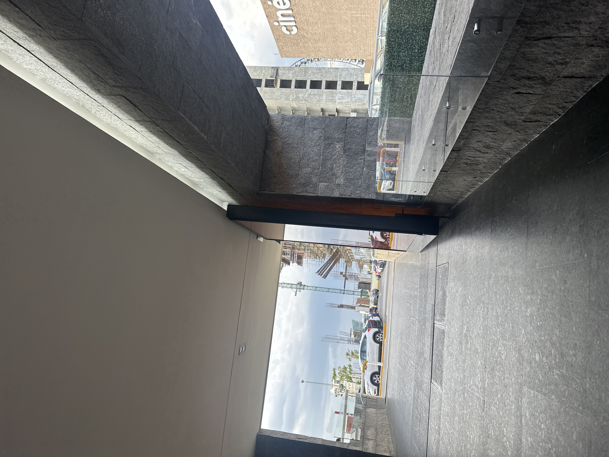 The image depicts an exterior view from within a covered walkway or tunnel leading out to an open area. The walkway has a dark gray, textured stone wall on the right and a light gray, possibly concrete, ceiling. A dark wooden door or frame is visible on the far right edge.
The opening of the walkway reveals a parking lot with several cars. In the background, construction is underway with visible cranes and scaffolding on unfinished buildings. To the right of the construction, a tan building with the word "ciné" in large white letters is partially visible, suggesting it is a cinema or part of a shopping complex. The sky is overcast.
Based on the presence of the "ciné" signage, which is common for Cinépolis cinemas, and the architectural style, the location appears to be a modern commercial development in Puebla, Mexico, possibly a shopping mall or entertainment complex like Explanada Puebla, which features a Cinépolis and ongoing construction projects.
