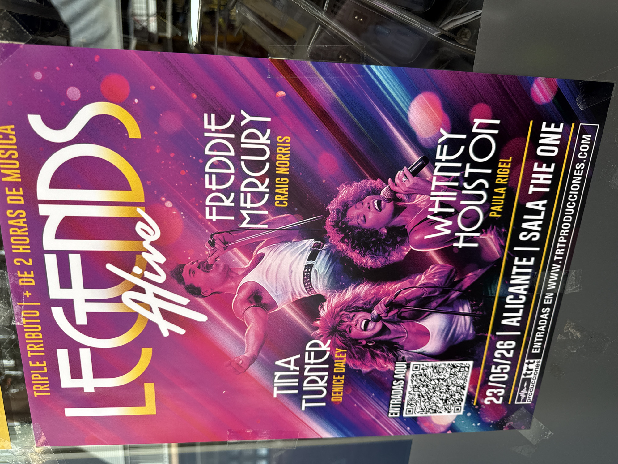 The image shows a promotional poster advertising "Legends Alive," a triple tribute concert lasting over 2 hours. The poster features images of performers impersonating Tina Turner (Denice Daley), Freddie Mercury (Craig Norris), and Whitney Houston (Paula Rigel). The concert is scheduled for May 23, 2026, and will take place in Alicante at Sala The One. A QR code labeled "ENTRADAS AQUI" (Tickets Here) and the website "WWW.TRTPRODUCCIONES.COM" are included for ticket information. The poster has a purple and blue color scheme with bokeh effects.