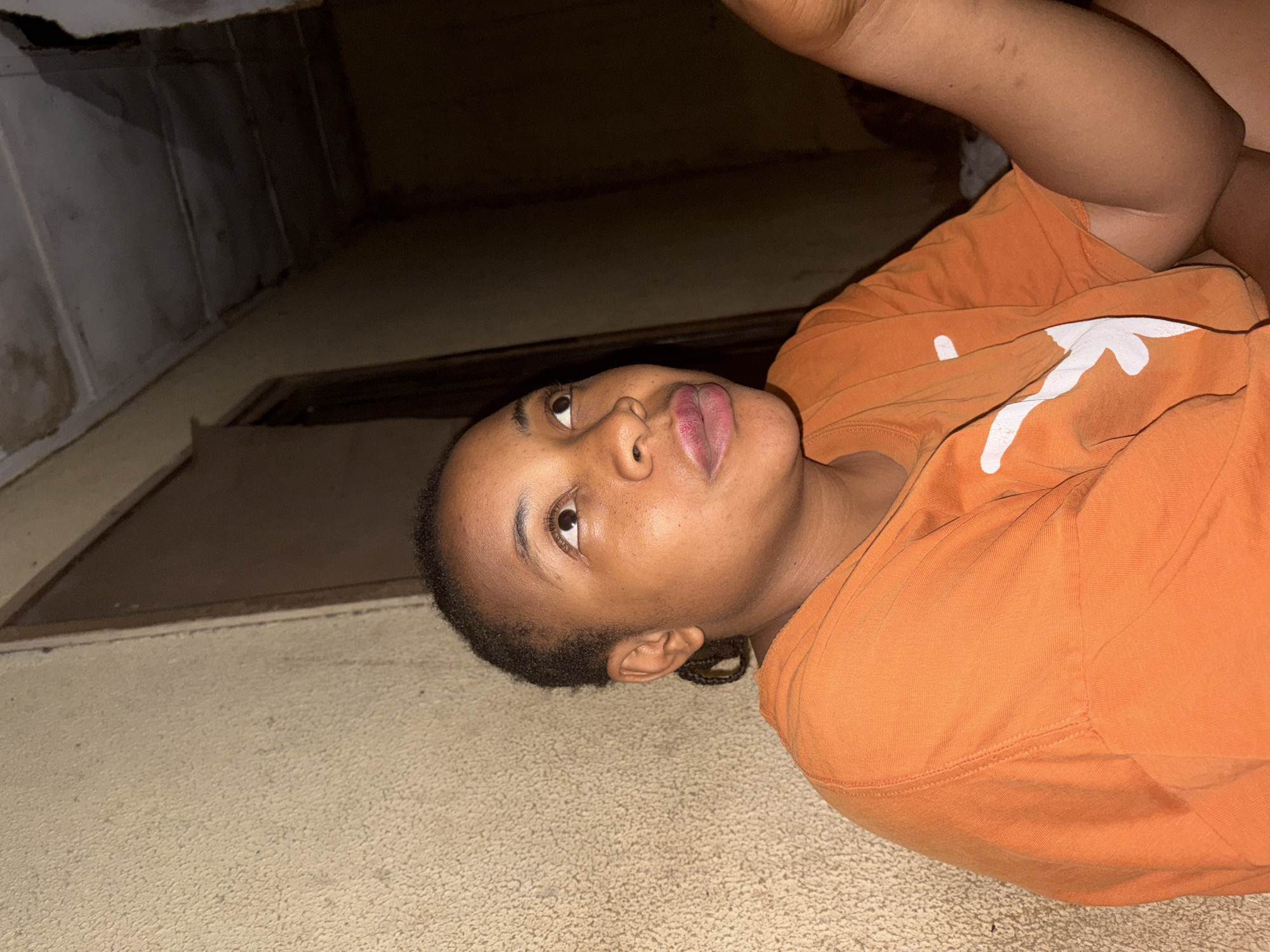 The image shows a young person with dark skin, likely female, captured from a rotated perspective, appearing to lie down or lean against a light-colored, textured surface, possibly carpet or a rough floor. The subject is wearing a bright orange t-shirt. Their head is turned, and they are looking upwards and slightly to the right (relative to the viewer if the image were properly oriented).
The person has short, dark hair, neatly braided, and visible eyelashes. Their lips are slightly parted, showing a reddish tint. One arm is visible, bent at the elbow, and the other is partially obscured, suggesting a relaxed pose.
A notable detail is the white logo on the front of the orange t-shirt, partially visible. This design strongly resembles the "Longhorn" or steer head symbol, commonly associated with the University of Texas at Austin.
The background is dark and indistinct, suggesting an indoor setting with low lighting. Structural elements such as walls or corners are faintly visible, contributing to the sense of an enclosed space. The dim illumination hints that the photo might have been taken at night or in a poorly lit room. The person's expression appears neutral, perhaps pensive or simply observant of something above them. No specific activity is occurring; it seems to be a posed or candid moment of rest. There are no direct visual cues in the image to confirm the provided location context of Jos, Nigeria.