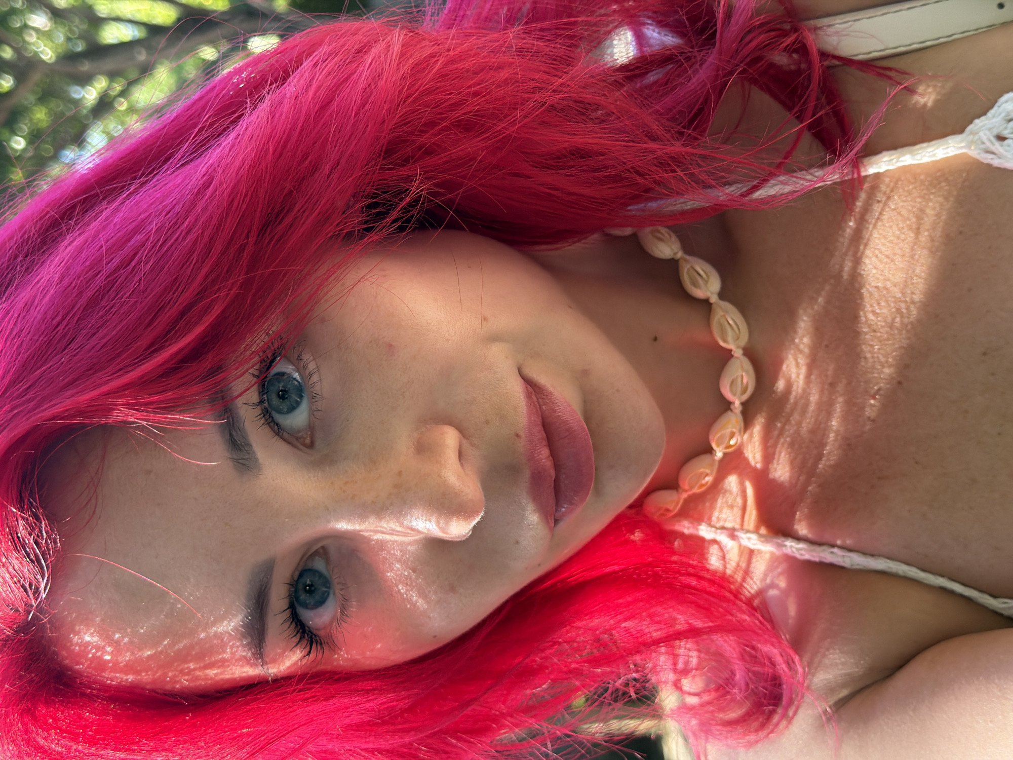 This is a close-up, tilted photograph of a young woman with vibrant, hot pink hair. Her hair cascades around her face and shoulders. She is wearing a white, crocheted top and a necklace made of natural, light-colored shells. The woman's face is turned towards the camera, and she has striking blue eyes, prominent eyelashes, and a light dusting of freckles across her nose and cheeks. Her lips are slightly parted, and she appears to be looking directly at the viewer with a gentle, perhaps playful, expression.
The lighting suggests it is daytime, with warm sunlight creating highlights on her skin and illuminating the texture of her skin. The background is softly blurred, showing out-of-focus green foliage and hints of dappled sunlight, suggesting an outdoor setting, possibly a garden or natural environment. There are no visible text elements in the image. The overall impression is one of natural beauty and a relaxed, possibly summery or tropical atmosphere, fitting with the provided location context of Kuta, Indonesia.