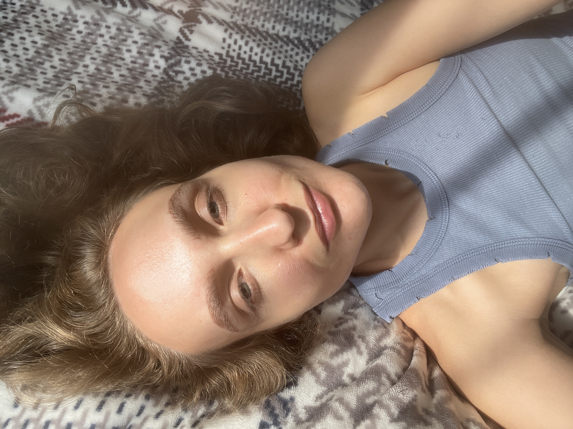 A young woman with light brown wavy hair lies on her back on a patterned blanket. She is wearing a light blue ribbed tank top. Sunlight casts shadows across her face and chest. The image appears to be a selfie taken from above. There is no discernible location in the city of Sharkan, Russia.