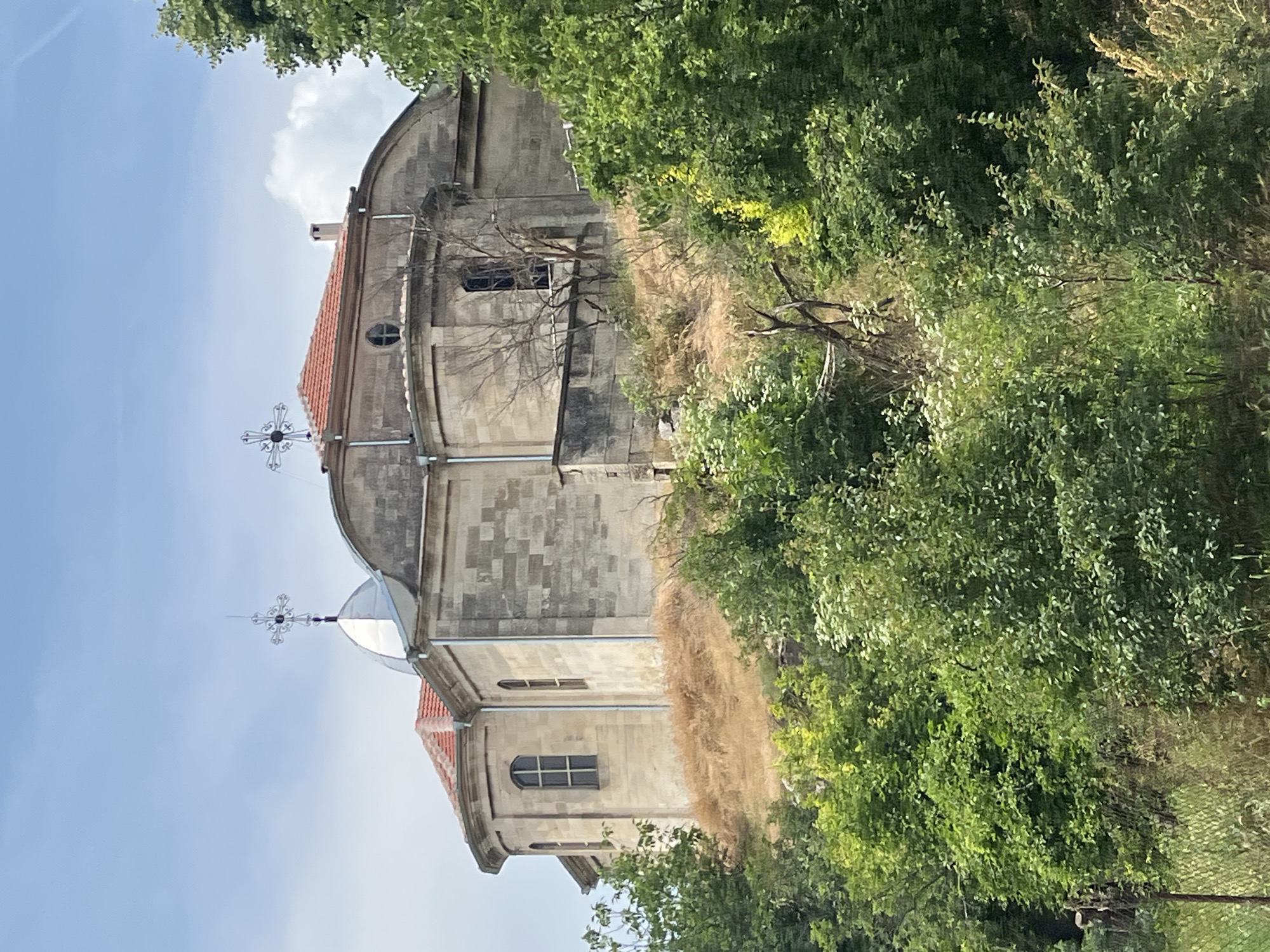 An elevated stone church or monastery, located on a hill in Basarbovo, Bulgaria, is depicted under a partly cloudy blue sky. The building features light-colored, possibly limestone, block construction with reddish-brown terracotta tile roofs. A prominent, reflective silver-colored dome or cupola, topped with an ornate dark metal cross, stands on the left side of the structure. A second, smaller ornate dark metal cross is visible on a terracotta-tiled roof section to the right. The building also displays multiple arched windows, one circular window, and a small rectangular chimney stack. Rain gutters are visible along its vertical edges. The immediate ground around the structure on the hill is covered in dry, light brown grass and sparse, parched vegetation. The lower and foreground sections of the image are densely populated with various shades of lush green deciduous trees and bushes. Some bare tree branches and skeletal vegetation are interspersed within the green foliage and on the dry hillside. White cumulus clouds are visible in the blue sky, particularly towards the right side of the frame. No individuals are present in the scene.