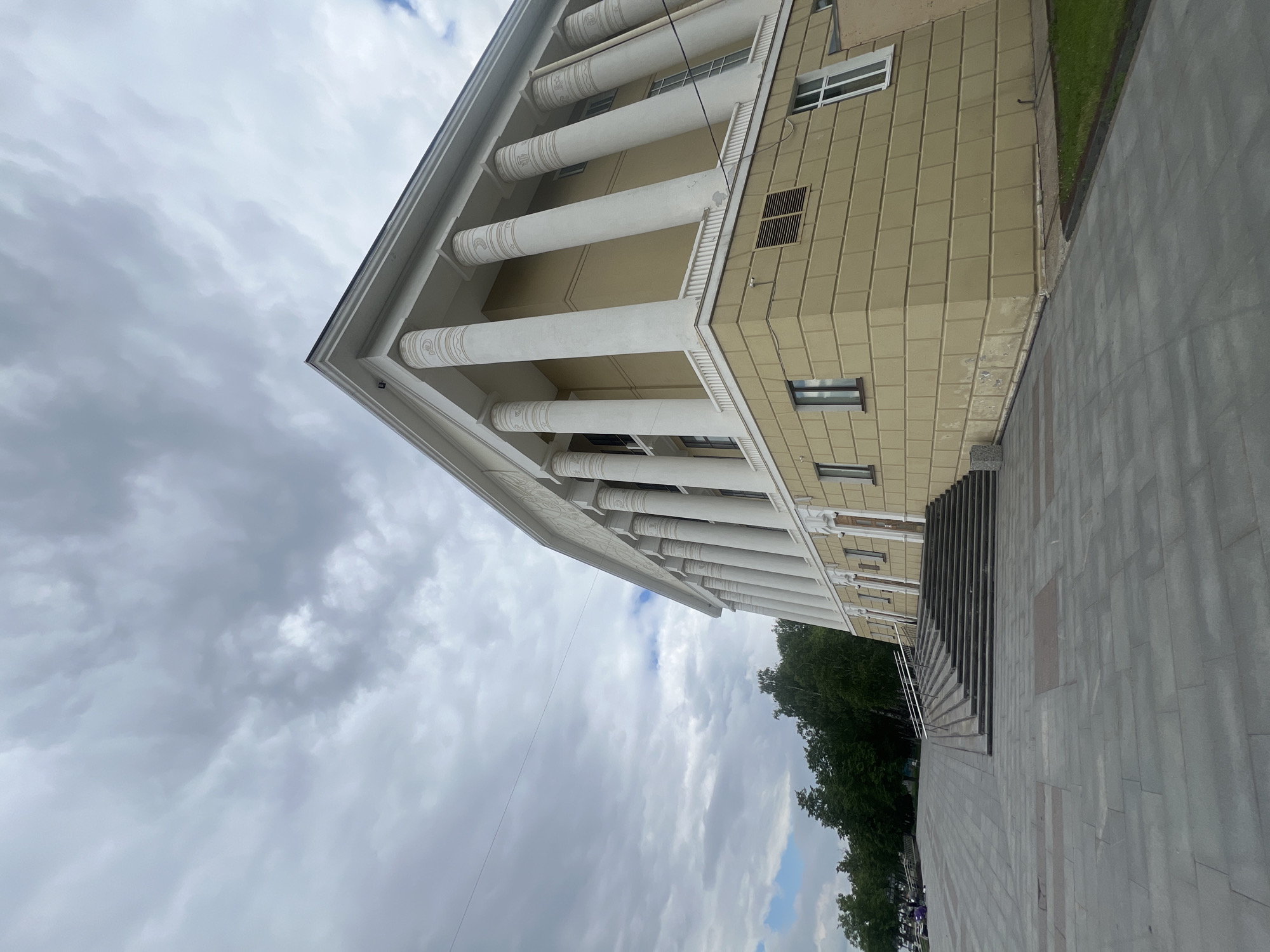 The image displays a grand, neoclassical-style building under an overcast sky in Votkinsk, Russia. The multi-story structure features a light yellow or tan facade with a rectangular, tiled pattern. A prominent architectural element is the series of tall, white columns supporting an overhang along the main front. These columns exhibit intricate relief patterns on their lower sections, adding to the building's ornate appearance. Several windows, some with white frames, are visible on the facade.
In front of the building, a wide, paved plaza or sidewalk made of gray rectangular tiles extends into the foreground. A broad set of dark, multi-tiered stairs leads from this paved area up to the building's entrance. To the right of the paved area, a strip of vibrant green grass runs alongside the building, followed by a dense line of dark green trees that obscure the distant background.
The sky is heavily clouded with various shades of gray, suggesting an overcast or approaching stormy day. A thin, dark line, possibly a wire or cable, diagonally crosses the sky in the upper left portion of the image. No people or specific activities are visible, presenting a static architectural and urban landscape. There is no discernible text in the image. The overall impression is one of a formal, institutional building.