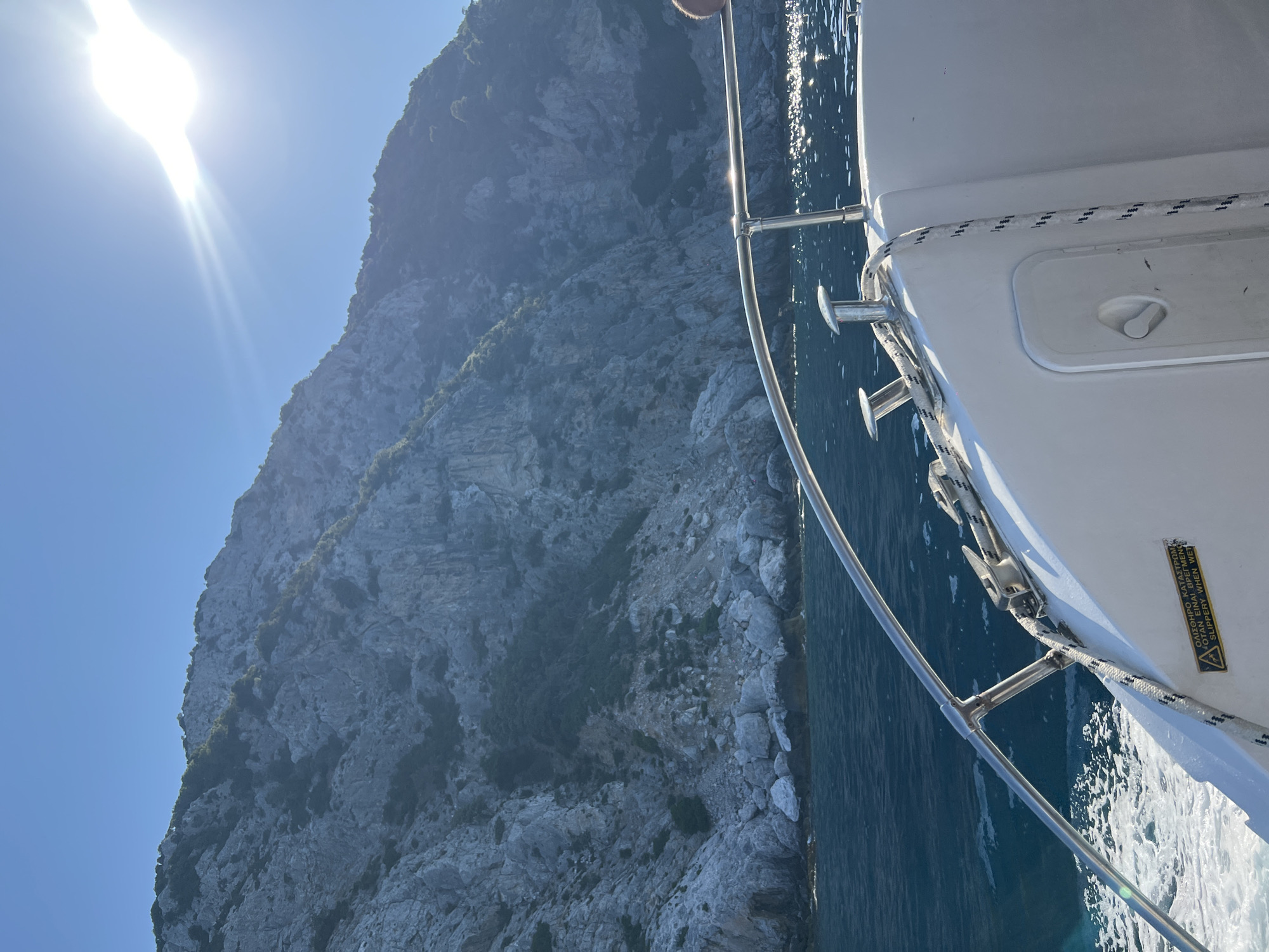 The image shows a view from a boat moving on the Aegean Sea towards the mountainous coastline of Skopelos, Greece. The water is a deep blue with white foam generated by the boat's movement. The sun shines brightly from the upper right of the frame, casting a glare on the water and partially obscuring the details of the rocky, vegetated mountainside. The boat's white hull and a metal railing are visible in the foreground. A coiled rope is secured to a cleat on the deck. A yellow warning label with text in Greek and English, indicating a slippery surface when wet, is attached to the side of the boat. The overall impression is one of a sunny day at sea with a dramatic coastal landscape.