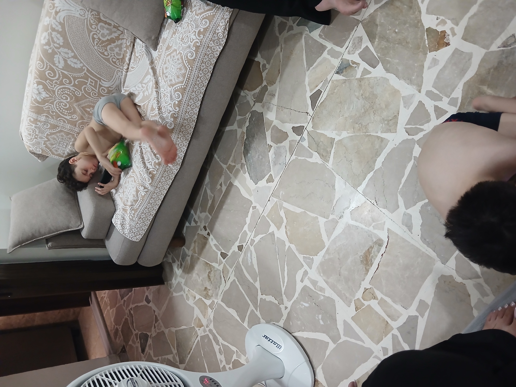 In a room with a terrazzo floor, a young boy is lying on his side on a sofa, holding a bag of chips and a black remote control. The sofa is covered with a patterned throw. To the left of the sofa, a white fan is positioned on the floor. In the foreground, another young child is on their knees facing away from the camera, with their head turned to the left. The room appears to be a living space within a home in Barza Municipality, Syria.