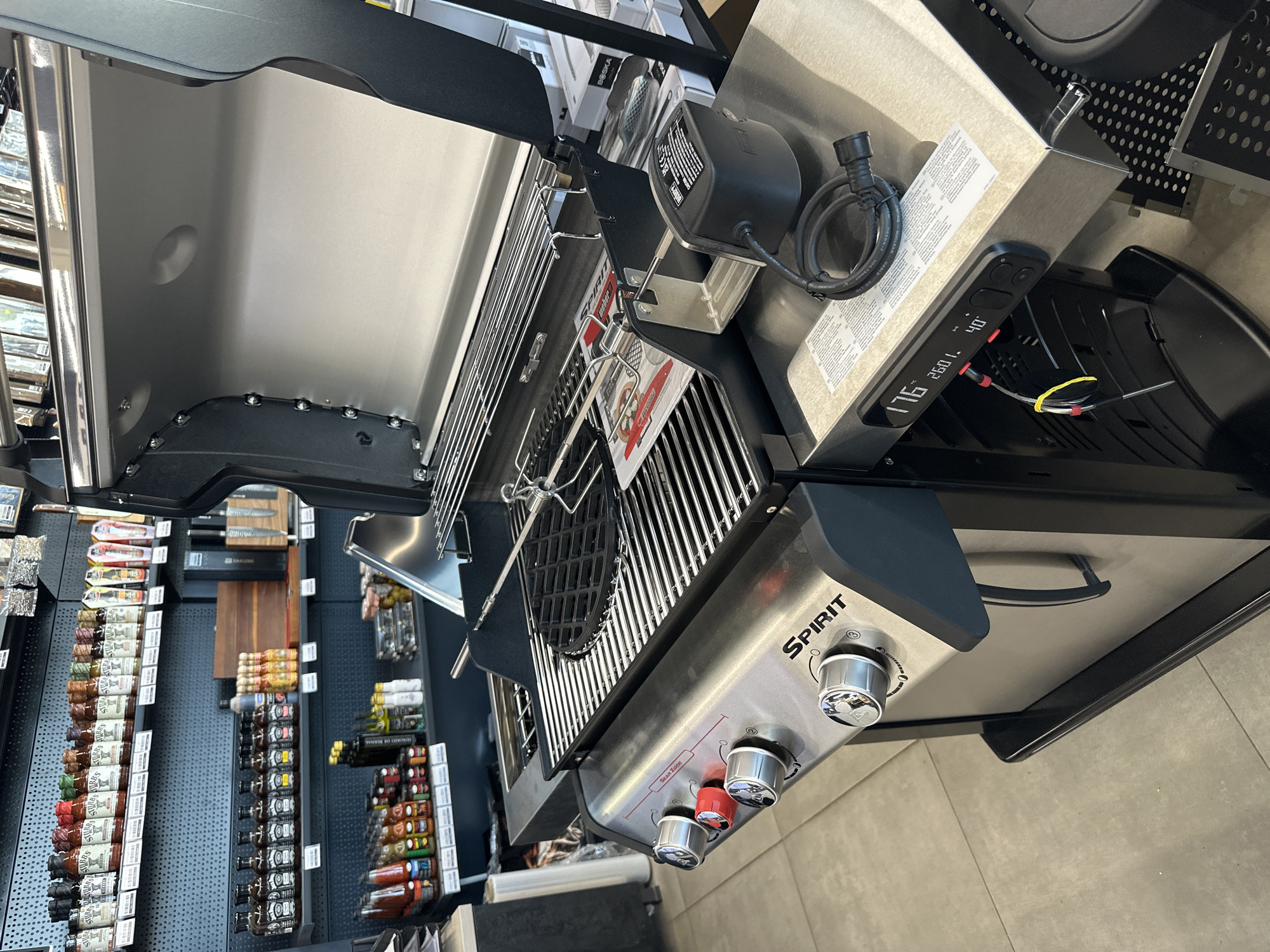 The image shows a stainless steel grill with the lid open, displaying multiple racks and a rotisserie. The grill has four control knobs on the front, one of which is red. A digital display panel with temperature readings is visible on the side. Various bottles of sauces and condiments are shelved in the background. The scene appears to be an indoor retail environment, possibly a kitchenware store or a specialty food shop. The floor is tiled. The grill is the primary subject of the image. There are no people visible. The location is not specified in the image or in the provided context.