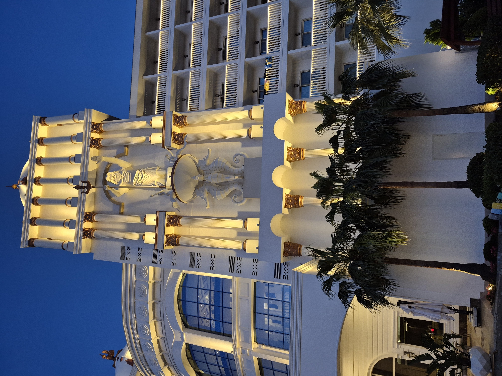 The image shows the exterior of a building in Alanya, Turkey at dusk. The building features white walls, multiple windows and balconies, and classical architectural elements, including columns and sculpted figures. The sky is a deep blue. Two palm trees are visible in front of the building.