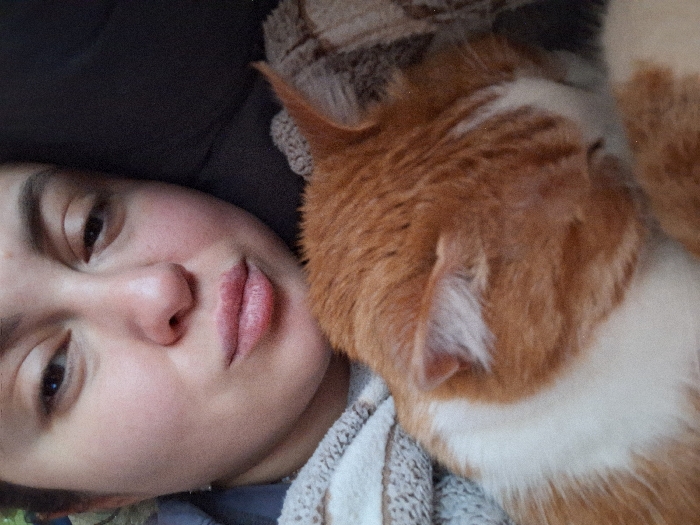 This close-up image depicts one adult female and one domestic cat in an indoor setting within Mikhalevo, Russia. The female's face, characterized by light skin and dark eyebrows, occupies the upper-left portion of the frame; her right eye is partially open and directed towards the viewer, her left eye is mostly closed, and her lips are slightly pursed. An orange and white cat is visible in the lower-right, primarily showing its back and a portion of its head, including its right ear, resting adjacent to the female. The female is positioned against a light-colored, striped textile, while the cat is situated against a darker, textured fabric.