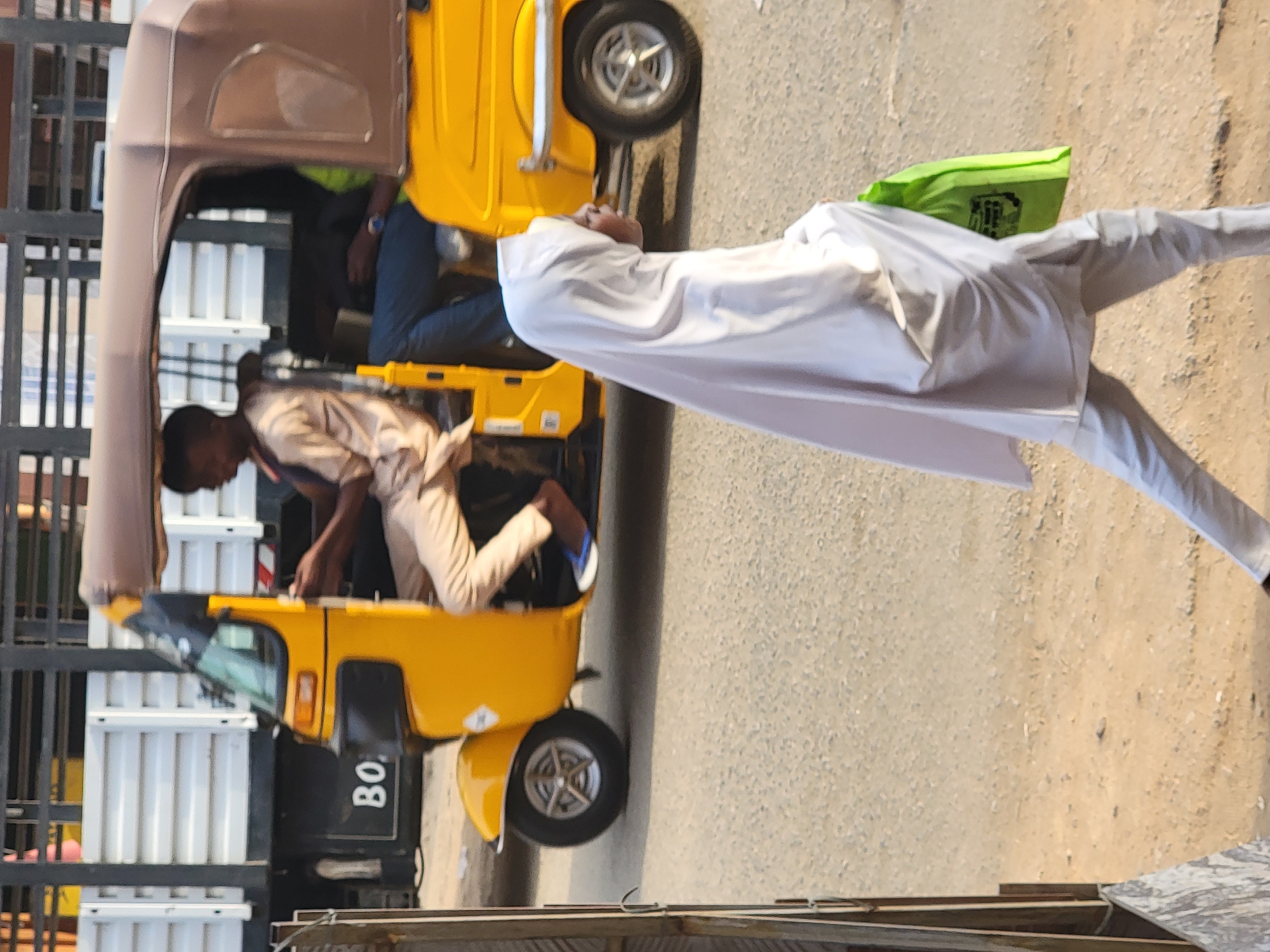 A partially sideways shot shows a yellow auto rickshaw, a three-wheeled vehicle, in an urban location in Maiburiji, Nigeria. One man is inside the rickshaw, appearing to drive, wearing a light brown shirt and dark pants. Another man, wearing a white gown and pants and carrying a bright green bag, is outside of the rickshaw, standing on the road. A building or structure with a black metal gate is to the left of the rickshaw. The road surface is visible.