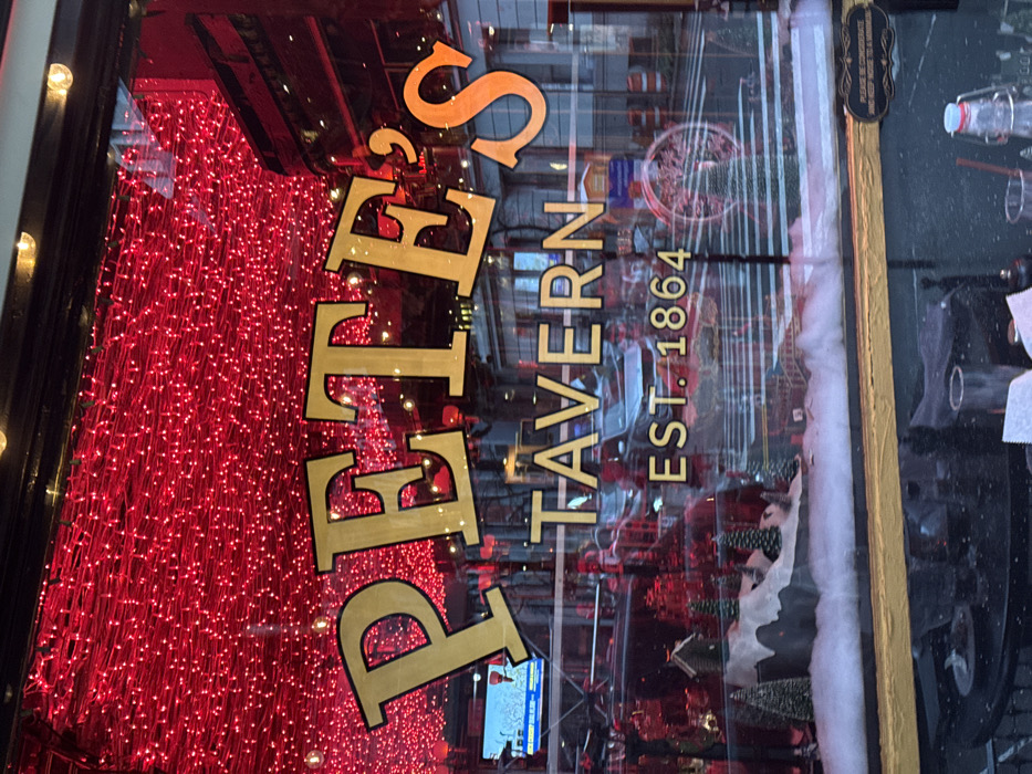 The image shows the exterior of "Pete's Tavern," established in 1864. The establishment's name is displayed in large, gold lettering on the window, reflected in the glass. The window is decorated with red string lights and a winter display featuring miniature trees and artificial snow. A television screen is visible inside, and the reflection shows the street.