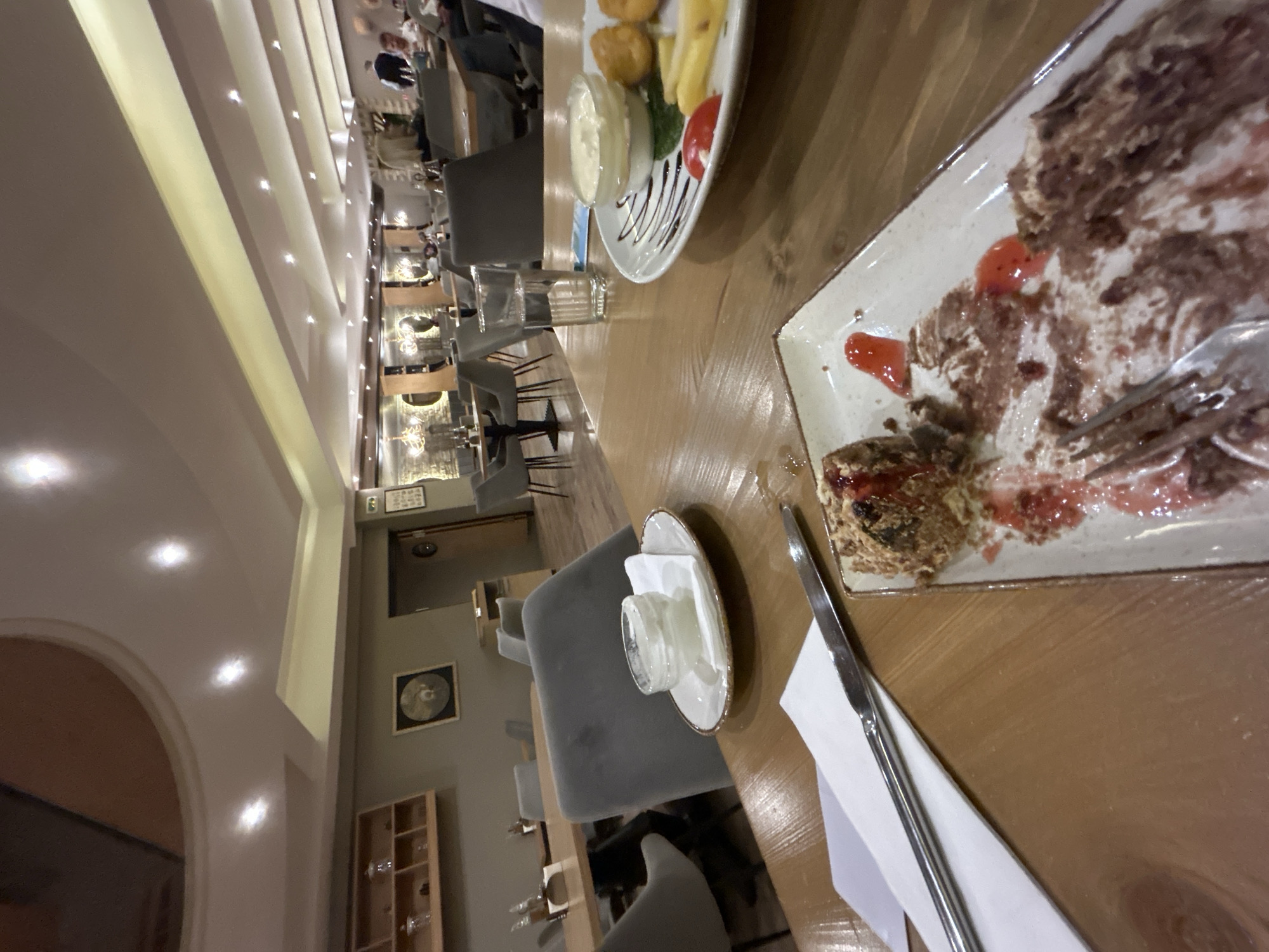 The image captures a long table in a restaurant, likely in Pamporovo, Bulgaria. The table is laden with remnants of a meal, including a plate with a partially eaten dessert, possibly a chocolate cake with cream and fruit sauce. Another plate contains fried items, such as fries and nuggets, accompanied by dipping sauce and tomato slices. A glass of water and a small dish with what appears to be a condiment or sauce sit nearby. Cutlery and a napkin are also present.
The restaurant is filled with chairs and tables. In the background, indistinct figures of people can be seen. The interior design includes archways, a shelf with glassware, and a framed circular design on the wall.