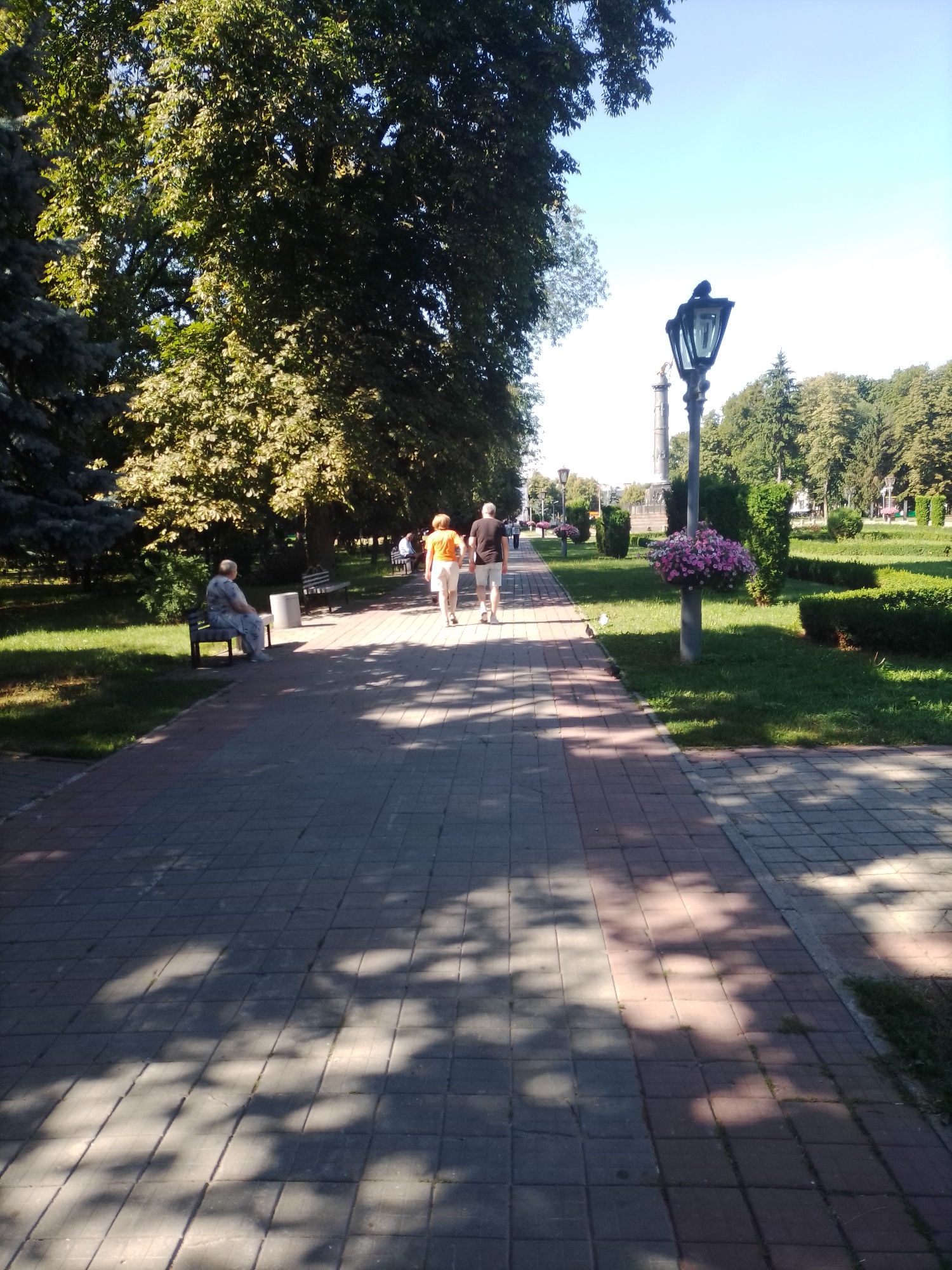 The image depicts a sun-dappled park walkway, likely in Poltava, Ukraine, on a clear day. Two individuals, a man and a woman, are walking away from the camera on the paved path, which is bordered by meticulously maintained green lawns and hedges. The woman wears an orange top and white pants, while the man is in a dark t-shirt and shorts. To the left of the path, an elderly person sits alone on a park bench, facing away from the viewer. Further down the walkway, a black ornate lamppost stands adorned with a hanging basket of pink flowers. The background is filled with lush, mature trees, suggesting a well-established park. The overall scene conveys a sense of peaceful outdoor activity.