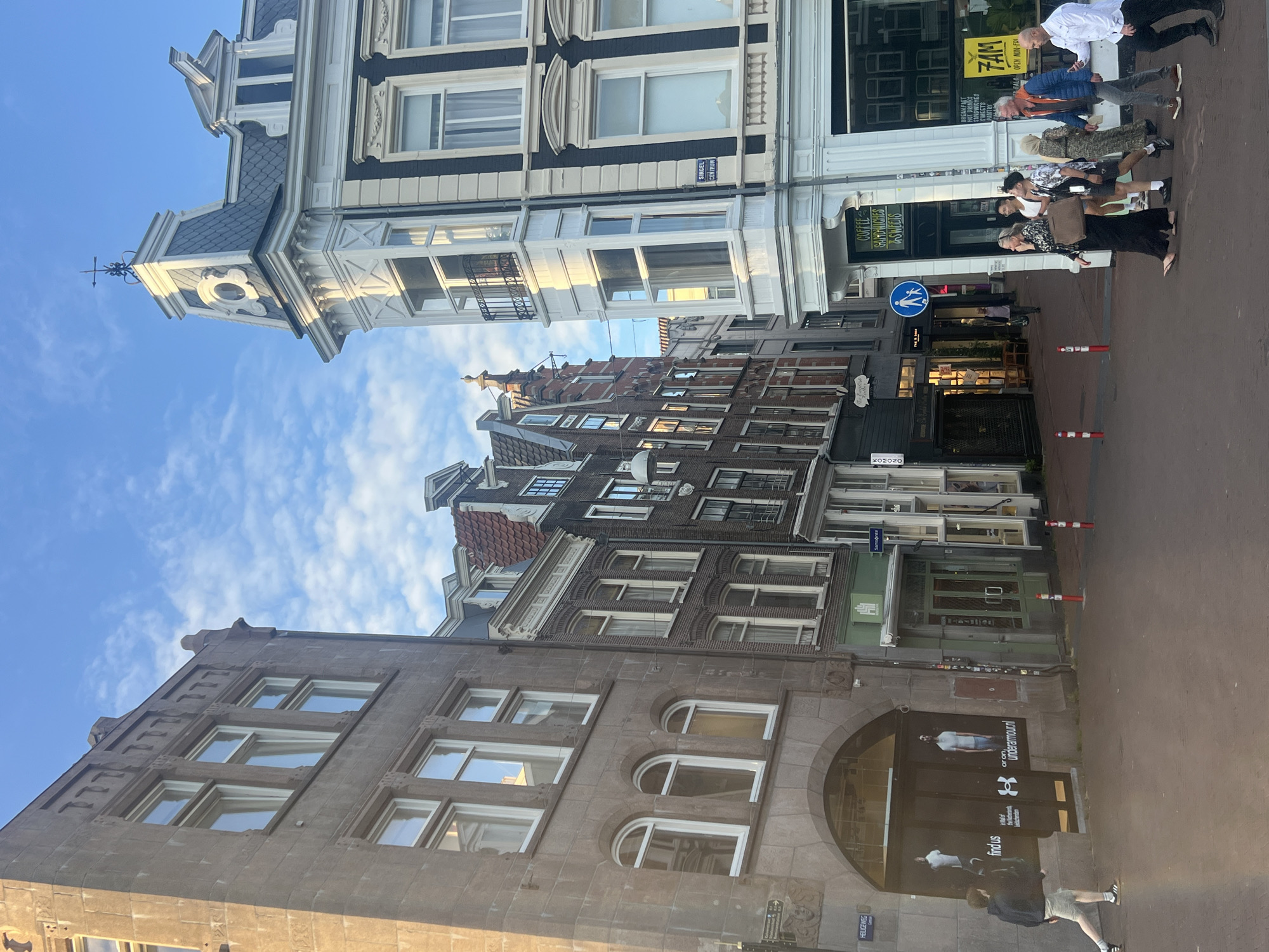 The image shows a street scene in Amsterdam, Netherlands, likely during the day due to the lighting. The architecture is characteristic of Amsterdam, with narrow buildings, gabled roofs, and large windows. On the left, a large building with a stone facade features arched entrances and windows. Several shops are visible along the street, including an Under Armour store with posters of models. Further down, there are shops with awnings and signs for "COFFEE SANDWICHES SWEETS" and "FAM".
Several people are walking along the street. On the left, a person in a dark hoodie and shorts walks away from the camera. In the center, a group of people are crossing the street, including women in dresses and a man in a white shirt and shorts. On the right, two men are walking together, one in a blue jacket and the other in a white shirt. The street is paved with brick. A pedestrian crossing sign is visible on a pole. The sky is blue with scattered clouds, suggesting fair weather. The overall impression is a busy urban environment with a mix of retail and residential buildings.