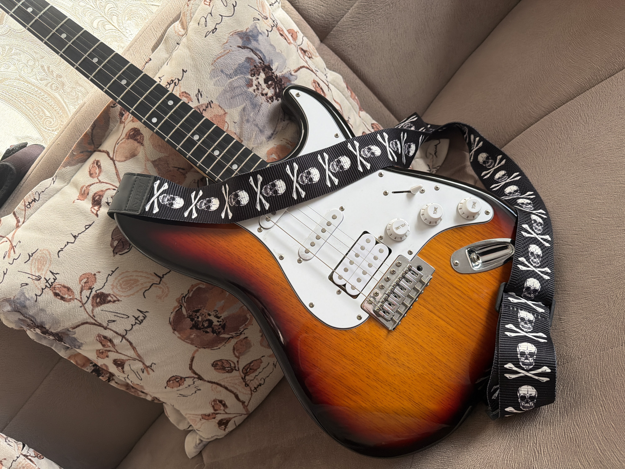 An electric guitar with a sunburst finish rests on a textured, neutral-colored sofa. The guitar is adorned with a black strap featuring a repeating pattern of white skulls and crossbones. The guitar neck is visible, with frets and fret markers clearly defined. On the body of the guitar, the white pickguard, chrome hardware, and control knobs are present. The guitar lies on a patterned cushion with floral motifs in muted colors. The scene is lit by natural light, suggesting it is daytime. There are no people visible in the image, and the setting appears to be indoors, likely a living room. The overall mood is still and casual. No visible text is present on the guitar or its accessories, other than what appears to be a small maker's mark on the strap's buckle.