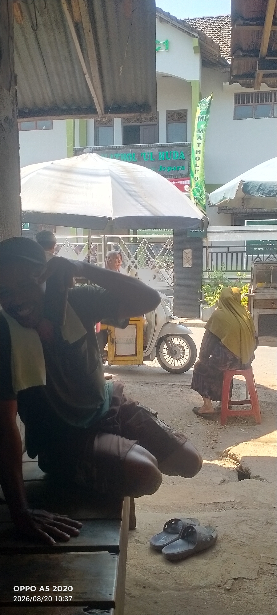 This is a medium shot photograph taken outdoors in Bugel, Indonesia, showing a street scene with several people and a building. The foreground is dominated by a person sitting on a wooden bench, partially obscured by shadows. This individual is wearing dark clothing and has a towel draped around their neck. To the right, a woman wearing a yellow hijab and a patterned dress is seated on a red plastic stool. Behind her, a white scooter with a yellow food cart attached is parked. In the background, a white building with a tiled roof is visible, featuring green signage that reads "MATHOL 'UL HUDA". The overall atmosphere suggests a warm, sunny day.