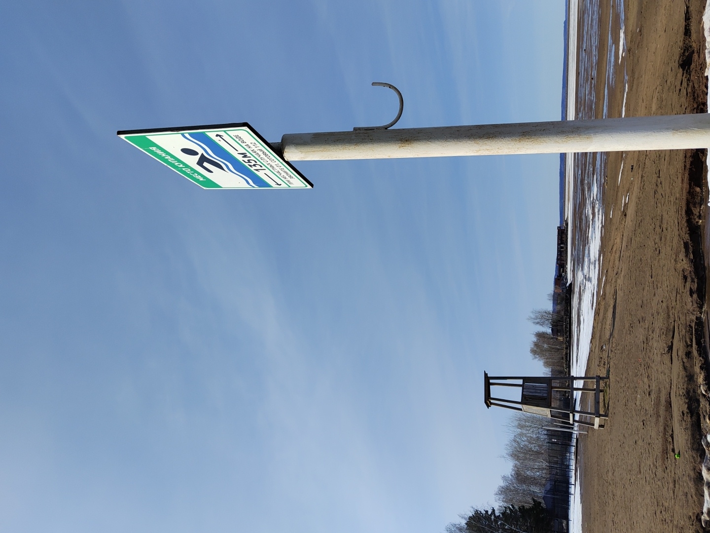 The image depicts a beach scene in the city of Chaikovsky, Russia. A sign indicating a swimming area, with Cyrillic text and an arrow pointing to "135m", is mounted on a pole. In the background, a lifeguard stand and a sandy beach are visible, with patches of snow. Trees line the area behind the beach. The sky is clear and blue. The image is rotated counter-clockwise.