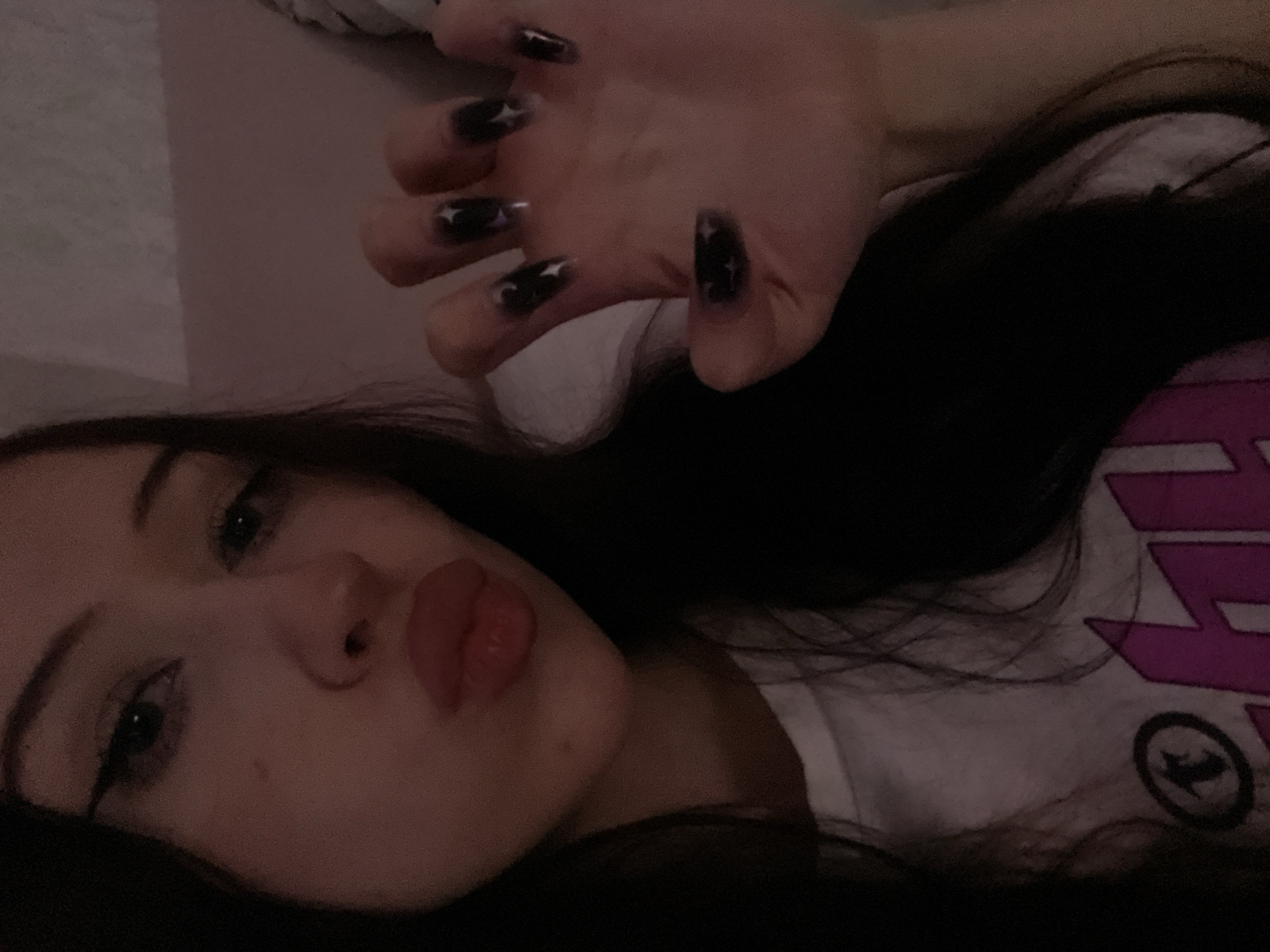 The media file is a close-up, selfie-style photograph of a young person lying down, with their face filling most of the frame. The person has long, dark hair and is wearing light-colored clothing with a design visible on the right side of the image. Their left hand is raised in the foreground, showcasing long, dark-colored fingernails adorned with star designs. The person's face is turned towards the camera, with their lips pursed in a "duck face" pose. Their eyes are open and looking towards the lens. The lighting is dim, suggesting an indoor setting, possibly at night or in a dimly lit room. The overall mood conveyed is casual and playful, likely taken in a personal or private setting. No specific location cues within Votkinsk, Russia are visible.